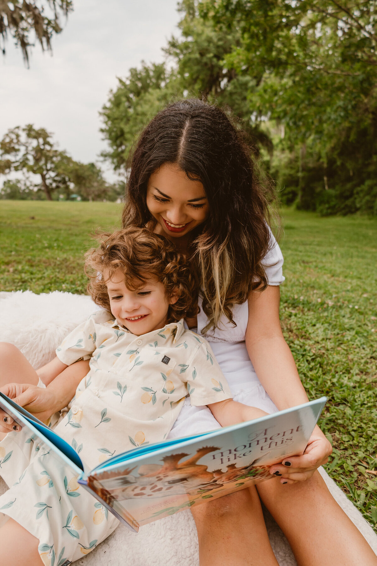 A candid moment of a mother and her young son reading a book together on a sunny day at Walter Hall Park. The pair is sitting on the grass, sharing smiles and enjoying a peaceful outdoor family session. 