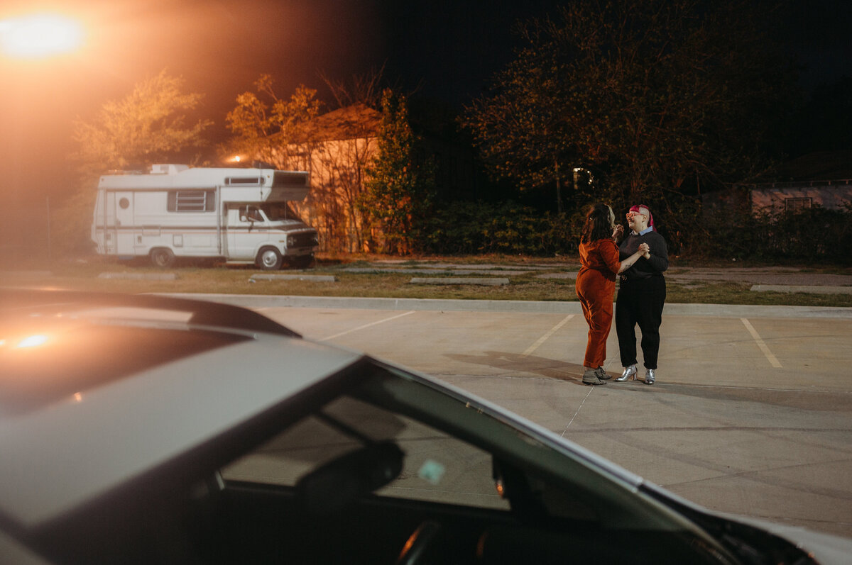 A couple holding each other in an empty parking lot at night, with a retro RV in the background