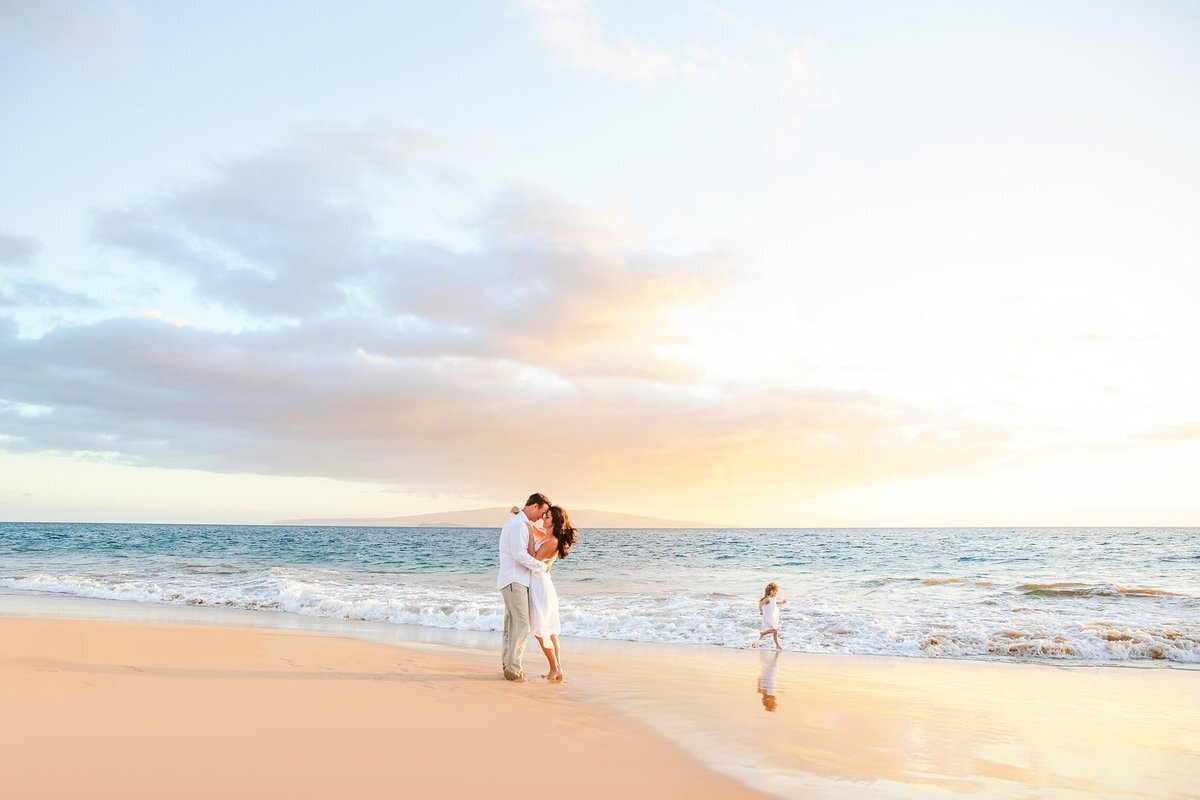 Sunset portrait on Maui with husband and wife kissing as little girl runs along the shoreline at the beach