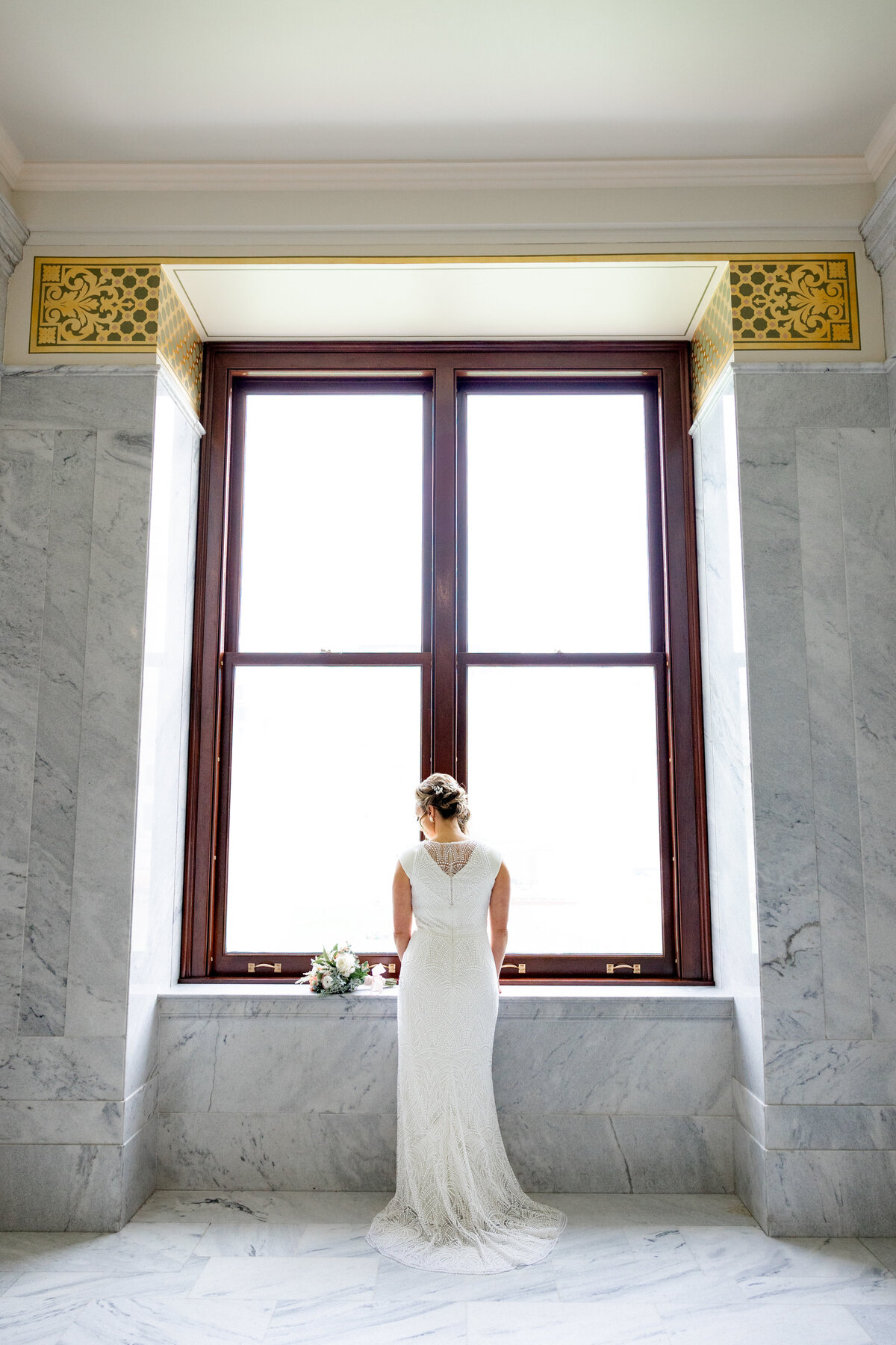 A bride stands by a large window in the Utah State Capitol, gazing out as her white lace gown flows behind her, with her bouquet resting nearby.