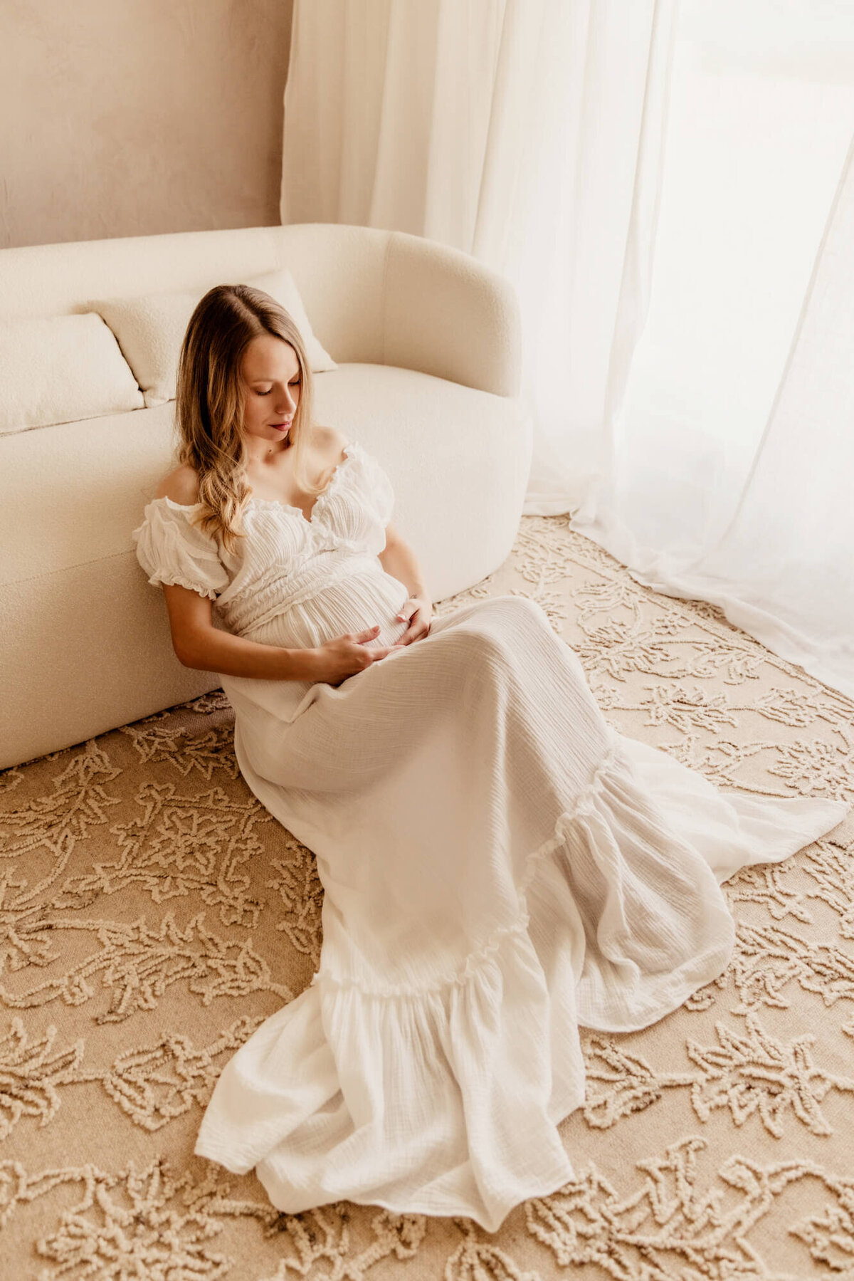 Mother holds her baby bump with both hands while sitting on a rug.