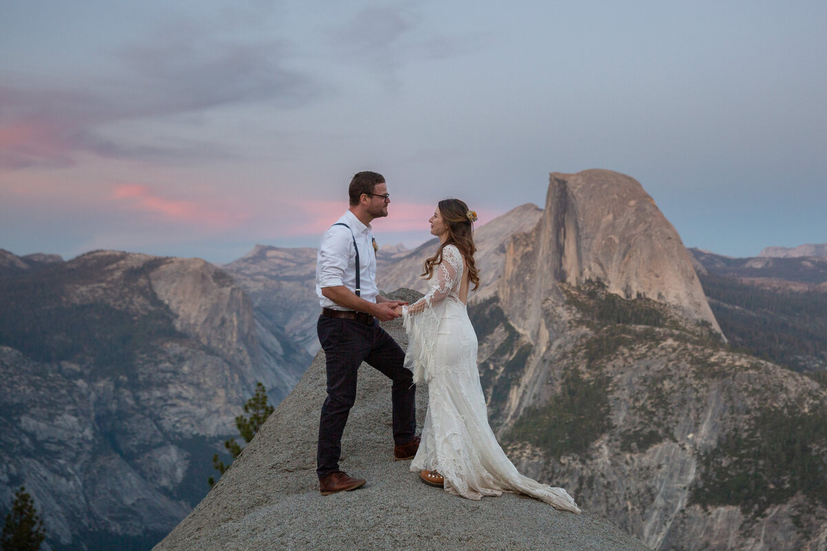 A bride and groom stand facing each other and holding hands on a rock in Yosemite National Park as the sunset and turns the sky pink behind them.