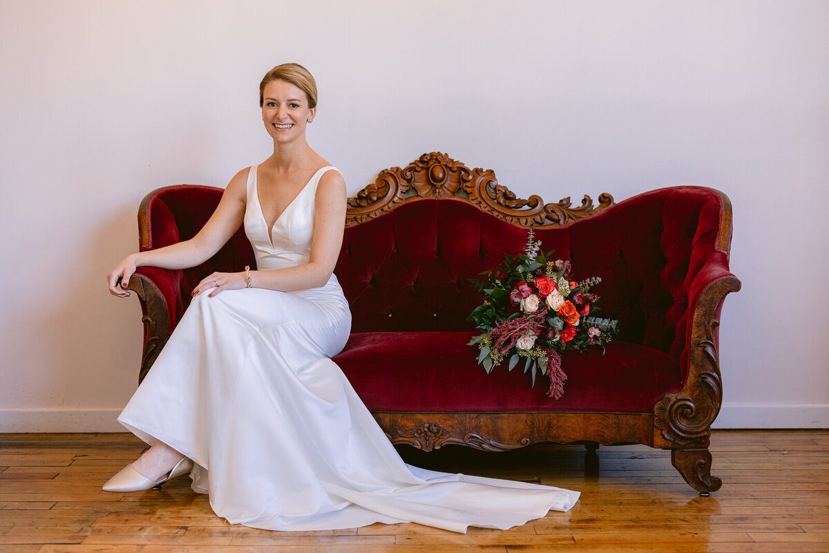 Wedding Photographer a bride sits on a red antique couch in her dress