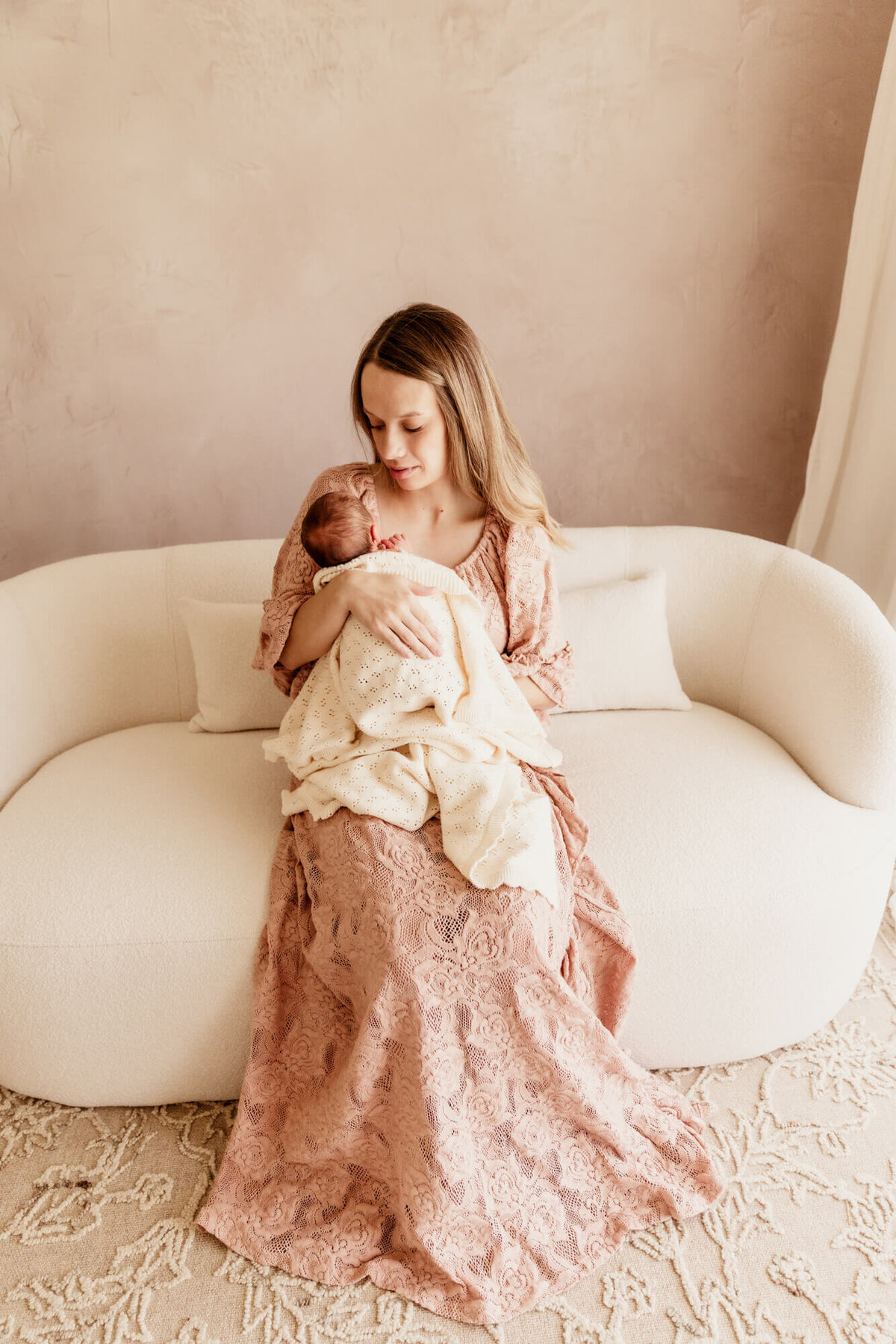 Woman wearing a pink lace dress holds her baby near her chest while looking down at her.