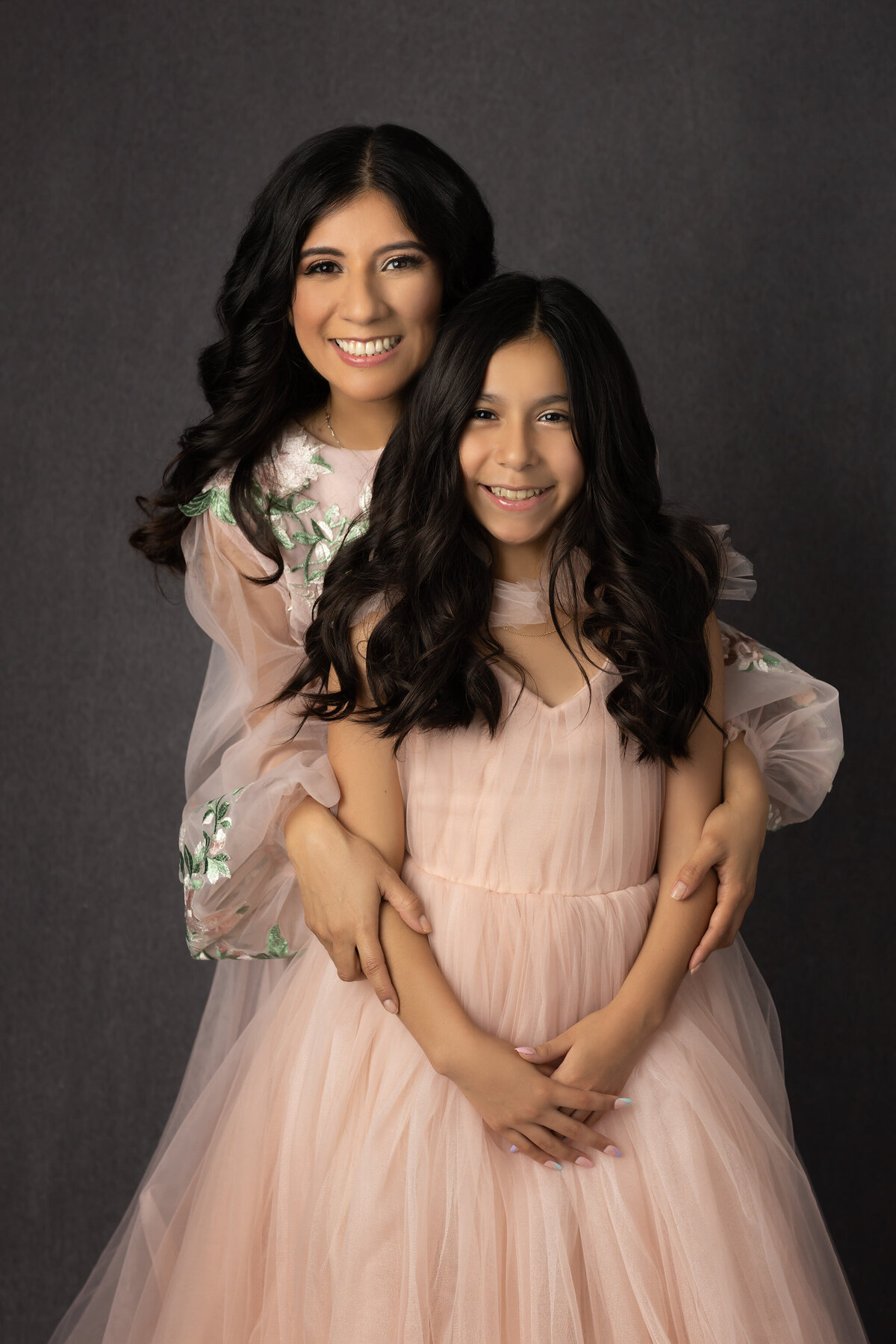 A Dallas family photographer captures a tender moment between a mother and daughter, both wearing matching soft pink dresses and smiling warmly.