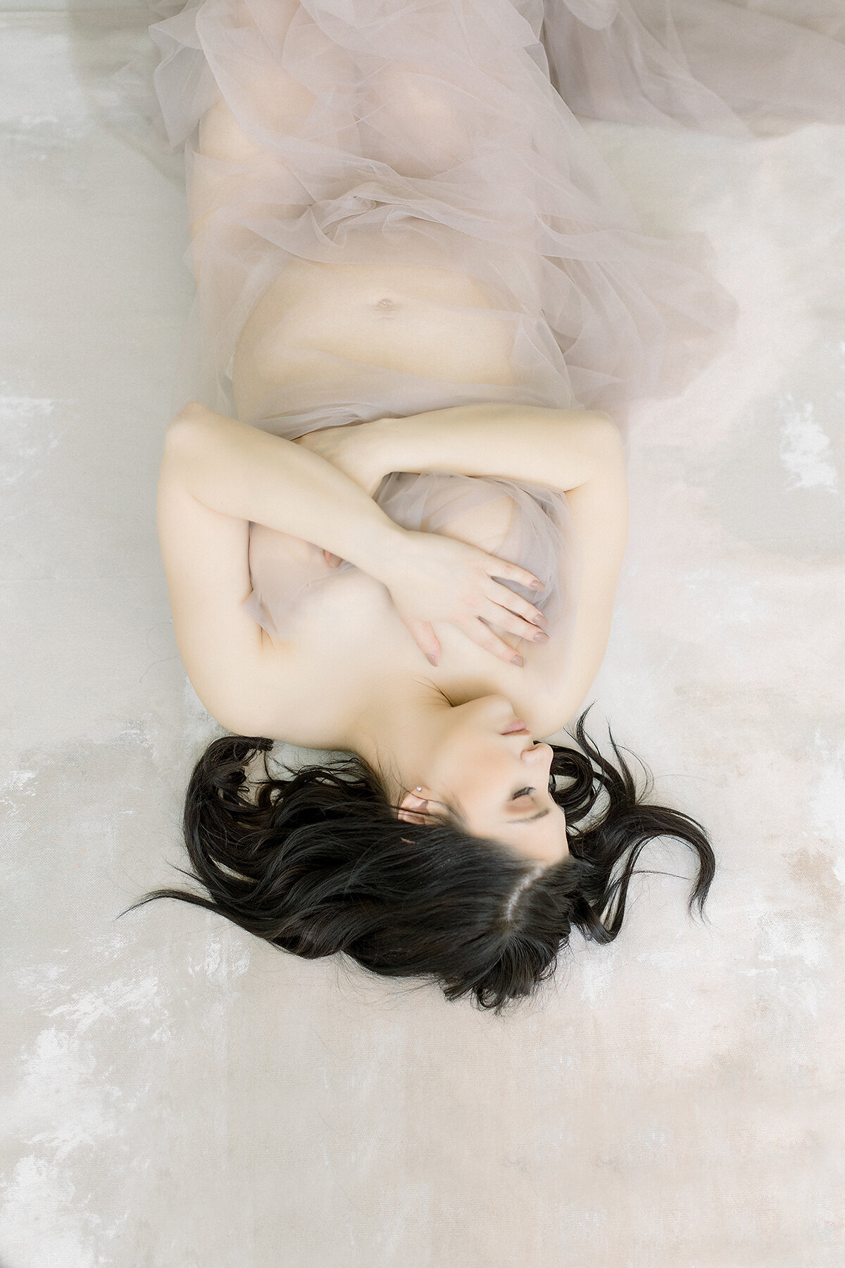 A light filled photo of an in studio maternity session of a pregnant DFW mother laying down on a bed draped in tulle as she is elegantly holding her chest and her belly.