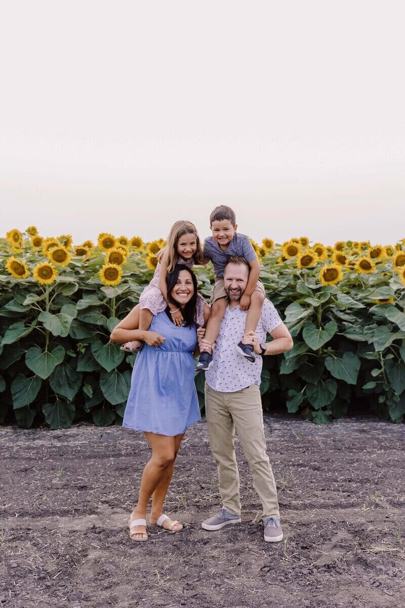 Family standing in a sunflower field, parents holding children on their shoulders while smiling at the camera.