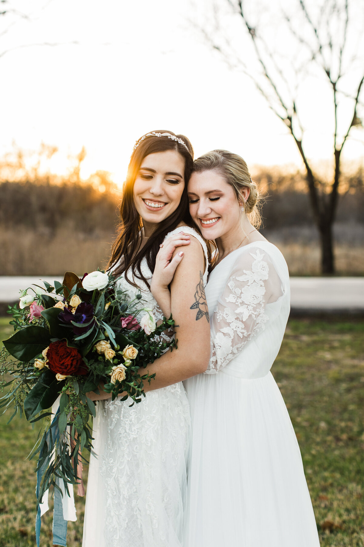 Portrait of two brides holding each other close while being the "big spoon" and "little spoon" on their wedding day in Dallas, Texas. The bride on the left is wearing a sleeveless white dress and detailed headband, is holding a large bouquet, and is the "little spoon." The bride on the right is wearing a long sleeve, detailed wedding dress, and is being the "big spoon."