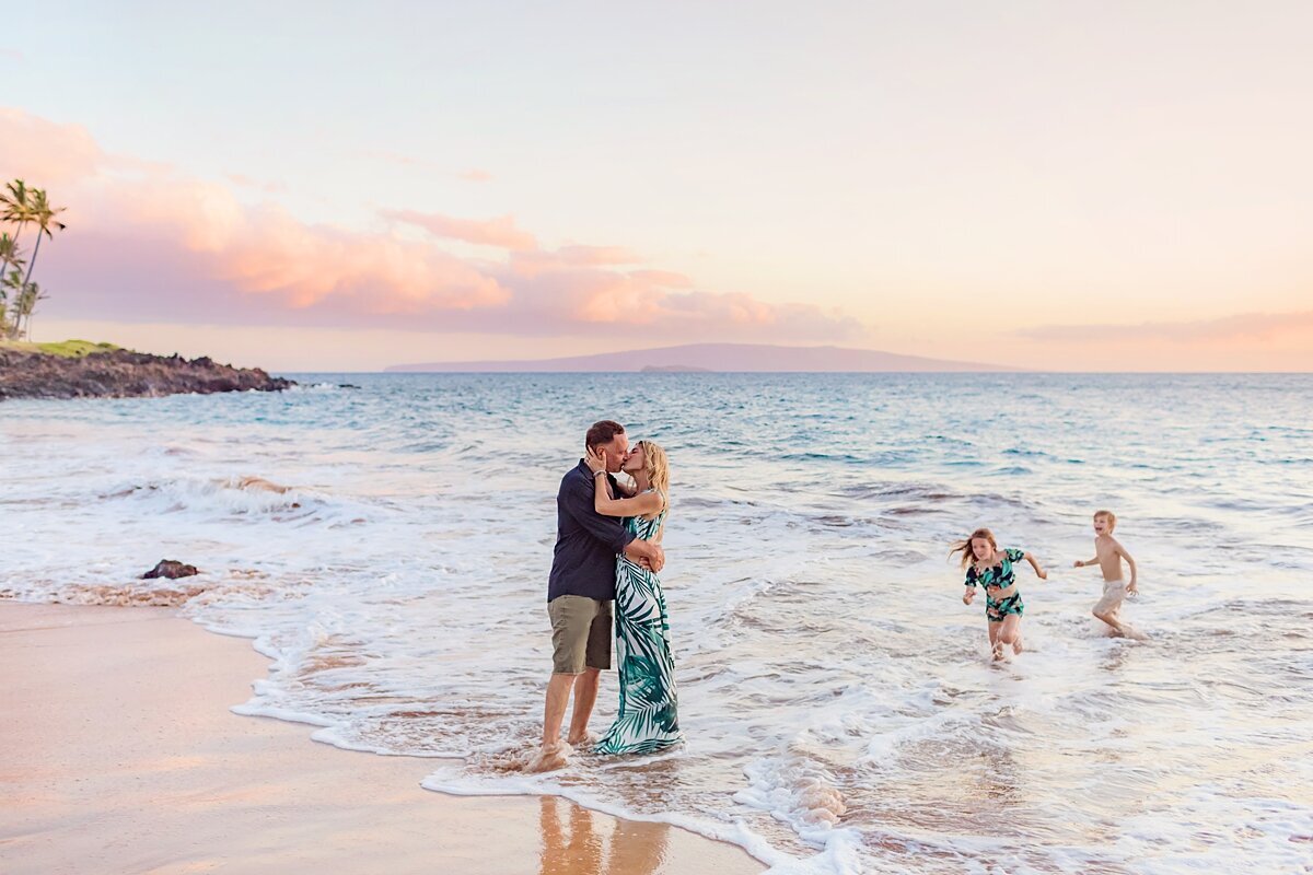 Candid family photography of husband and wife in green dress kissing next to the ocean as their two young kids run in the waves behind them