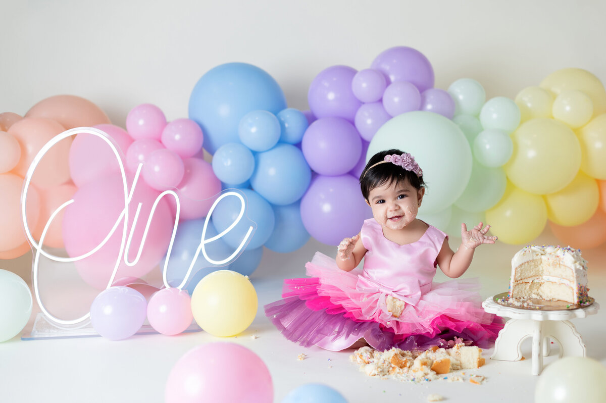 Baby girl laughs, covered in cake, rainbow balloons. Houston child photography.