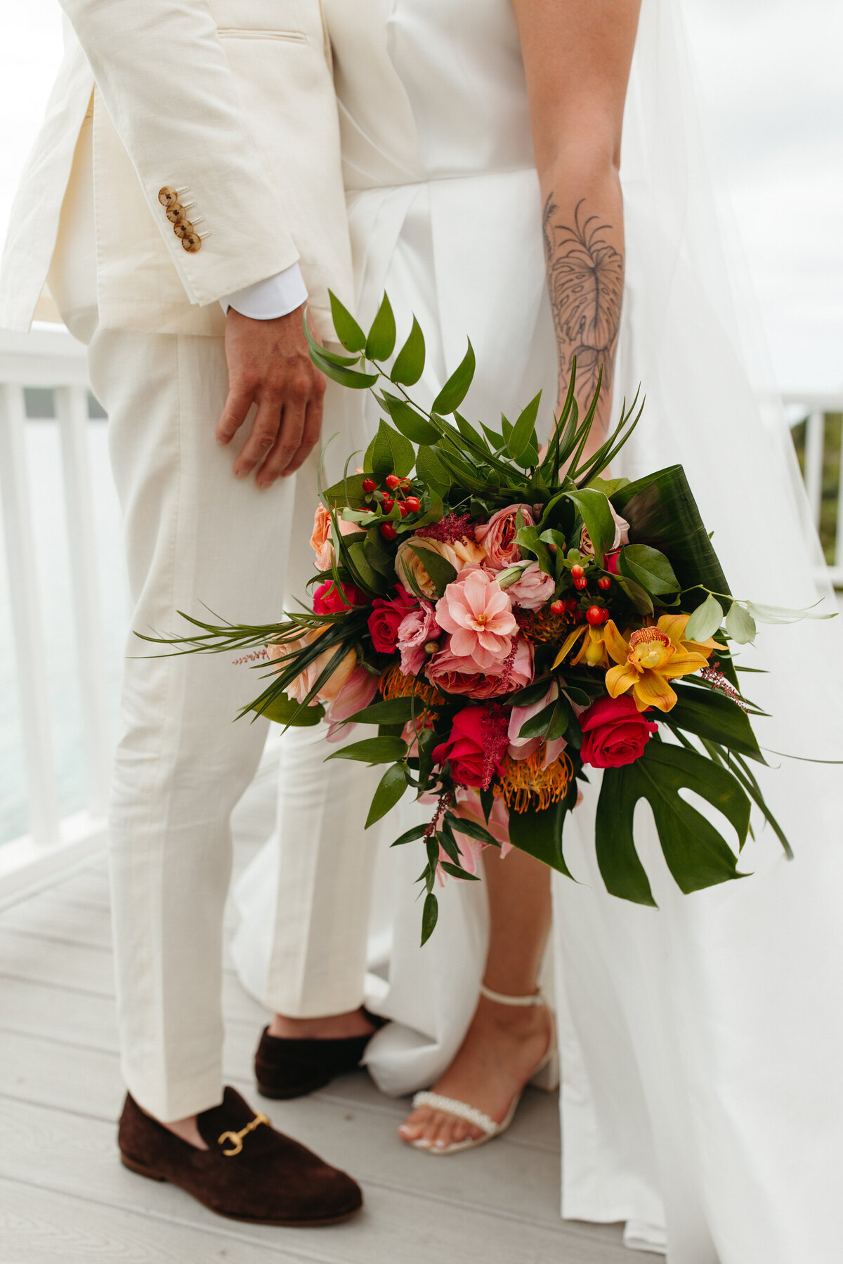 Close-up of a lush wedding bouquet with vibrant tropical flowers cradled by a person in a white suit next to a white-dressed partner