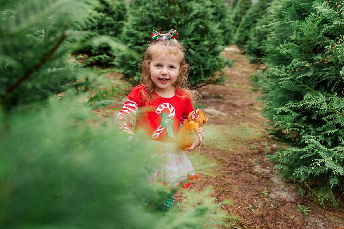 little girl wearing a christmas outfit playing behind a christmas tree at a christmas tree farm in Savannah, ga