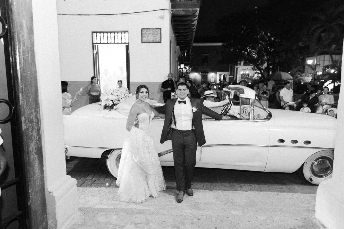 A timeless black-and-white photo of Victoria and Daniel leaving in a vintage car after their Cartagena destination wedding ceremony.