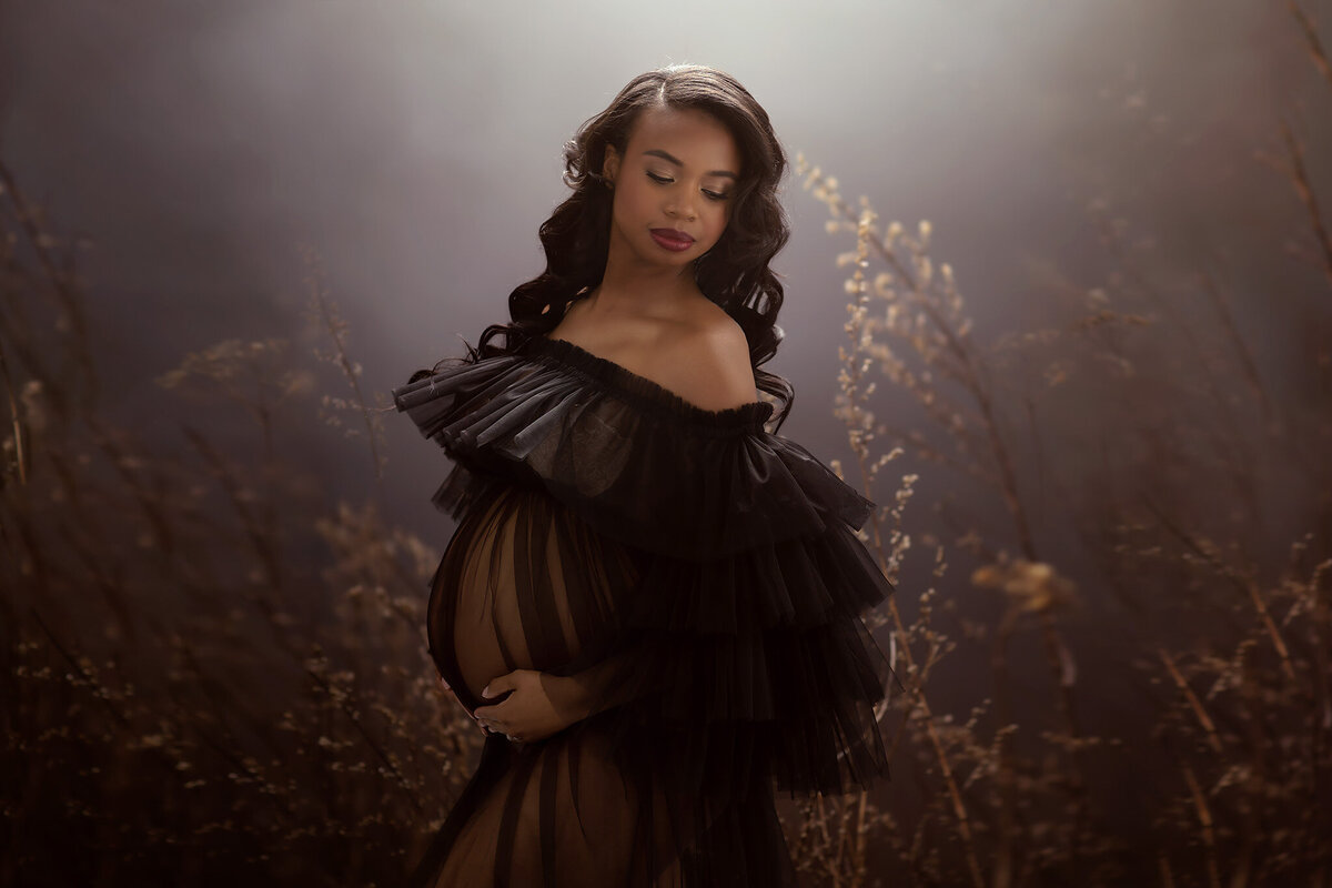 A Dallas maternity photographer captures a pregnant woman in a sheer black off-shoulder dress, standing in a field of dry grass. She cradles her belly, looking down tenderly, as soft, glowing light envelops her maternity moment.
