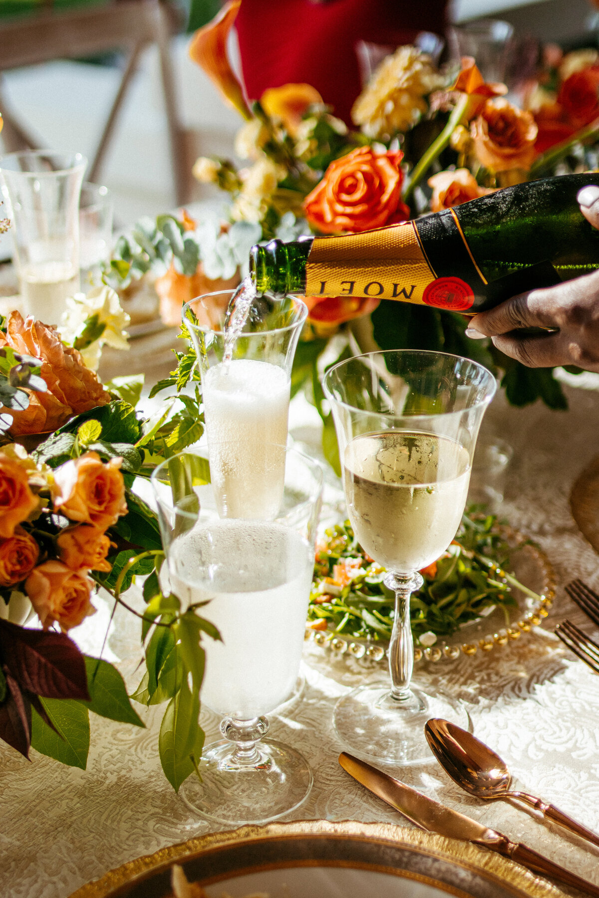 hand pouring moet champagne on a table set with mixed metals place settings drinking glasses and orange rose centerpieces