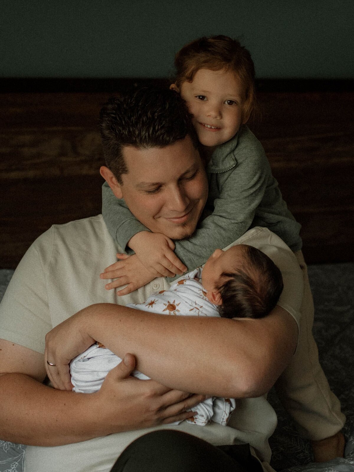 Father holding newborn while big sister wraps her arms around him, a heartfelt family moment from an in-home newborn session in Murrieta.
