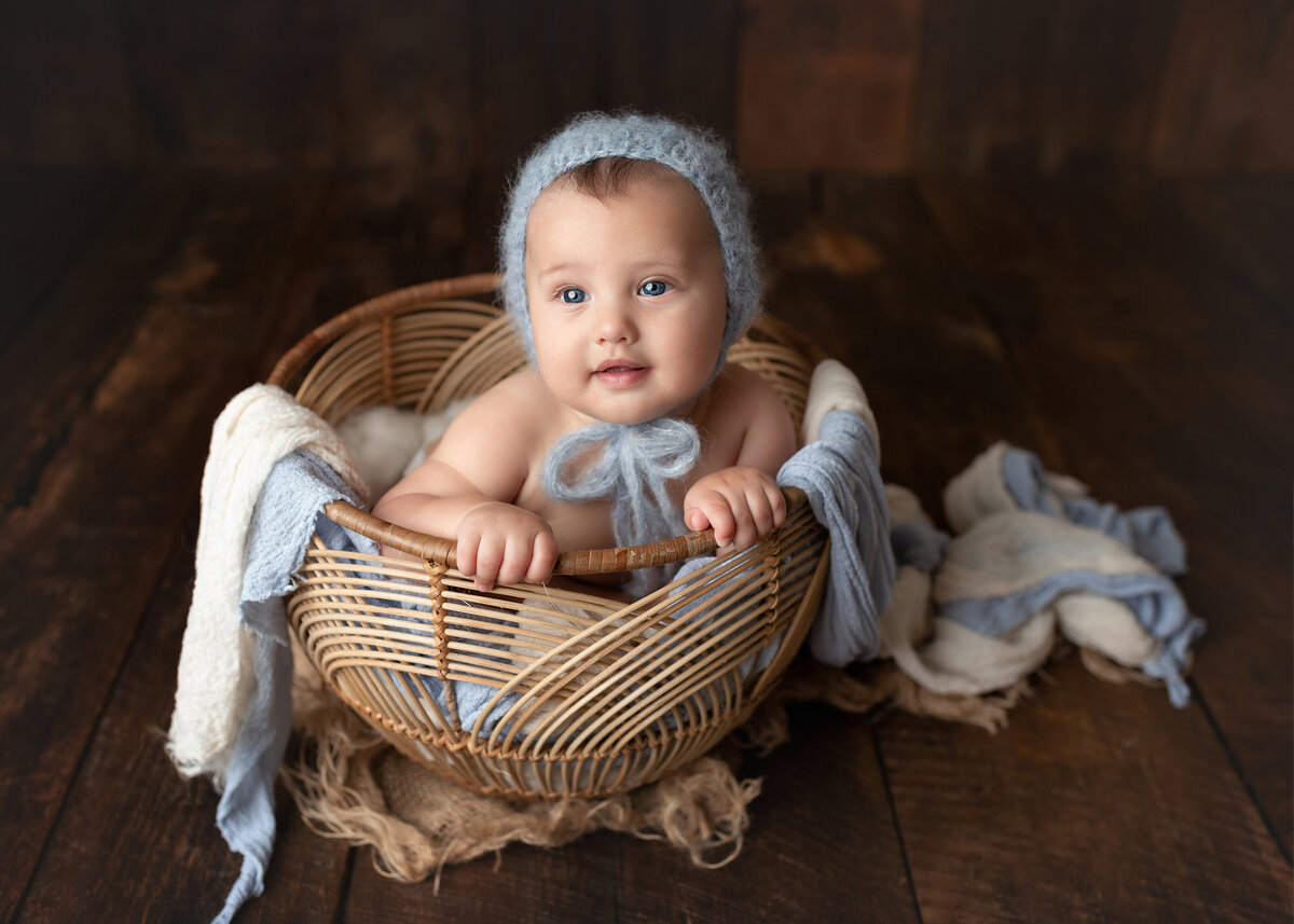 Baby boy 6-month milestone photoshoot. Baby boy is in a rattan basket that is draped with blue and white linen fabric. Baby's hands are clutching the edge of the basket and is wearing a blue knit bonnet.