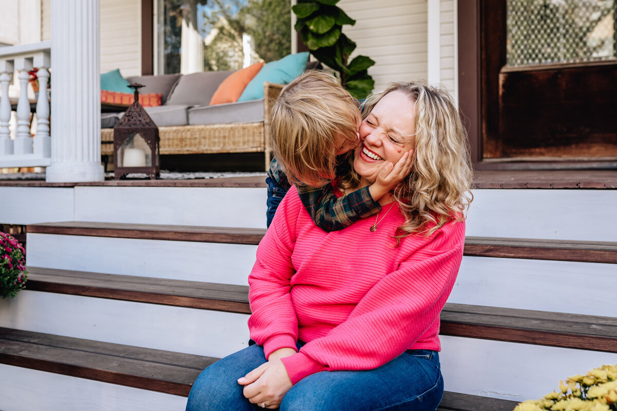 A mom sits laughing on a porch while her child kisses her cheek.