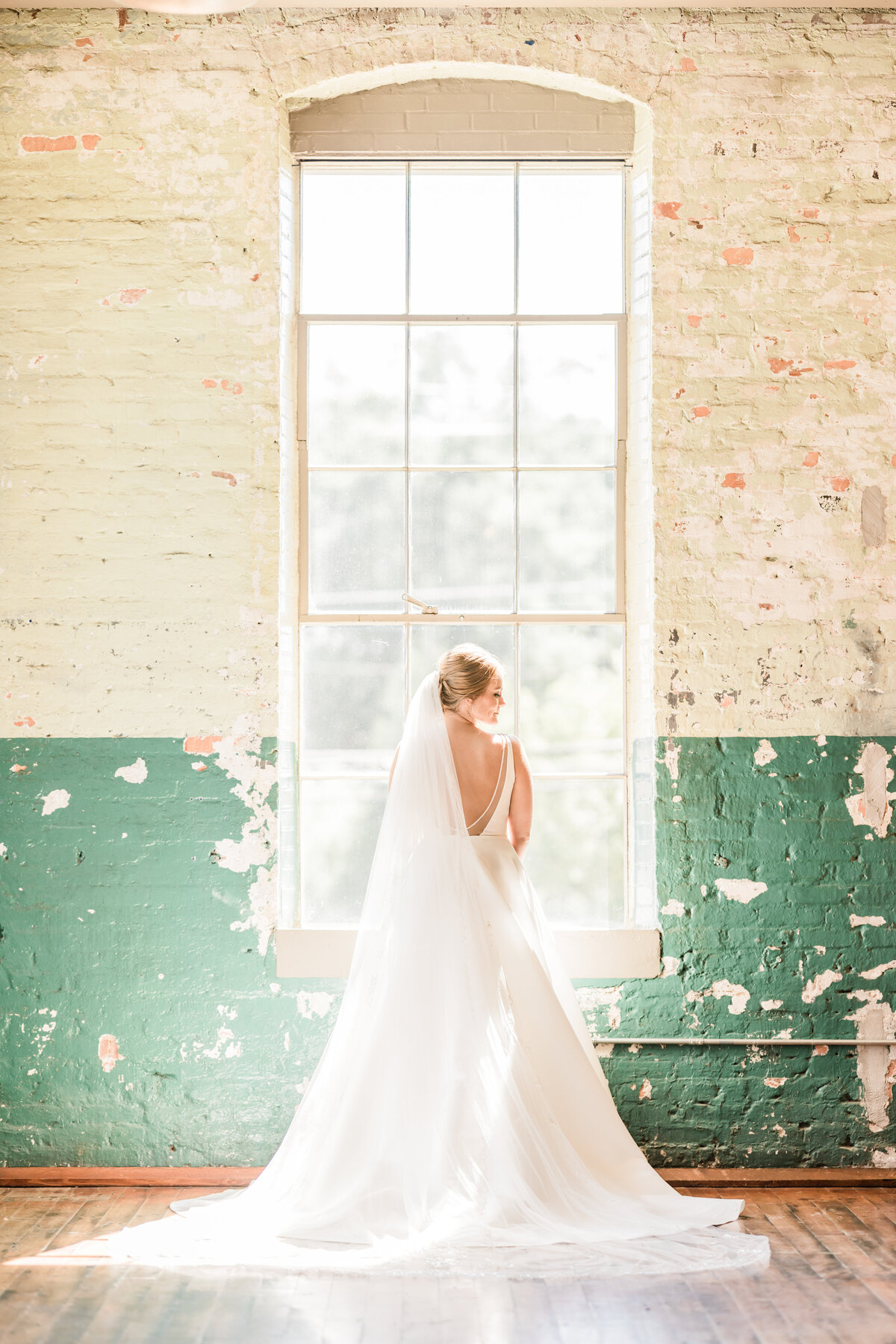 Bride looks out window next to textured walls of The Engine Room Wedding Venue in Monroe, GA. 