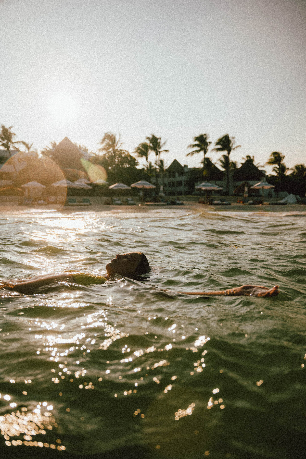 Person floats on their back in sunlit ocean waters near a tropical beach. Palm trees and beach umbrellas are visible in the background, with the sun casting a warm glow over the scene.