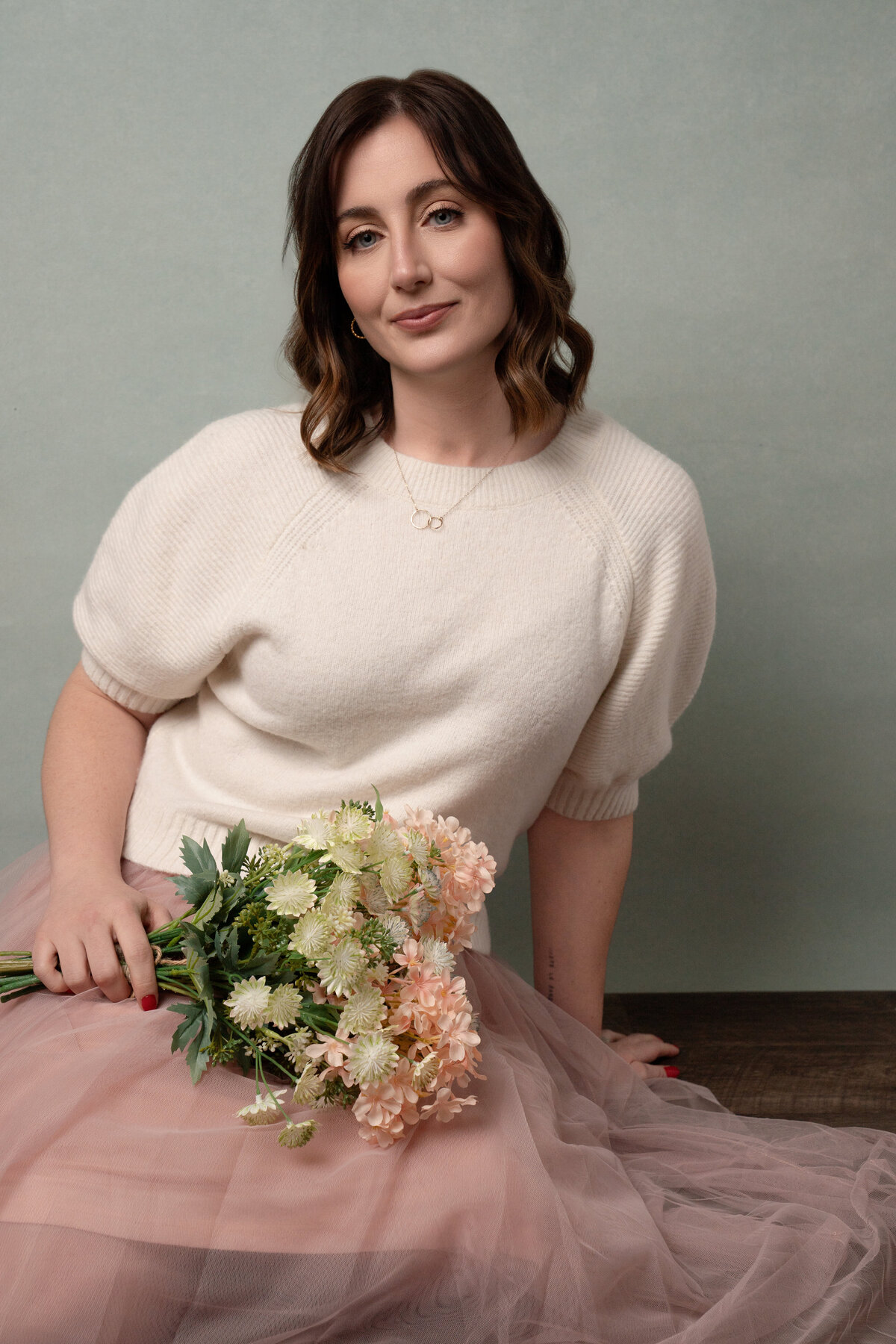 Woman sitting on floor holding bouquet of flowers wearing short sleeve off white sweater and pink tulle skirt that matches flowers.