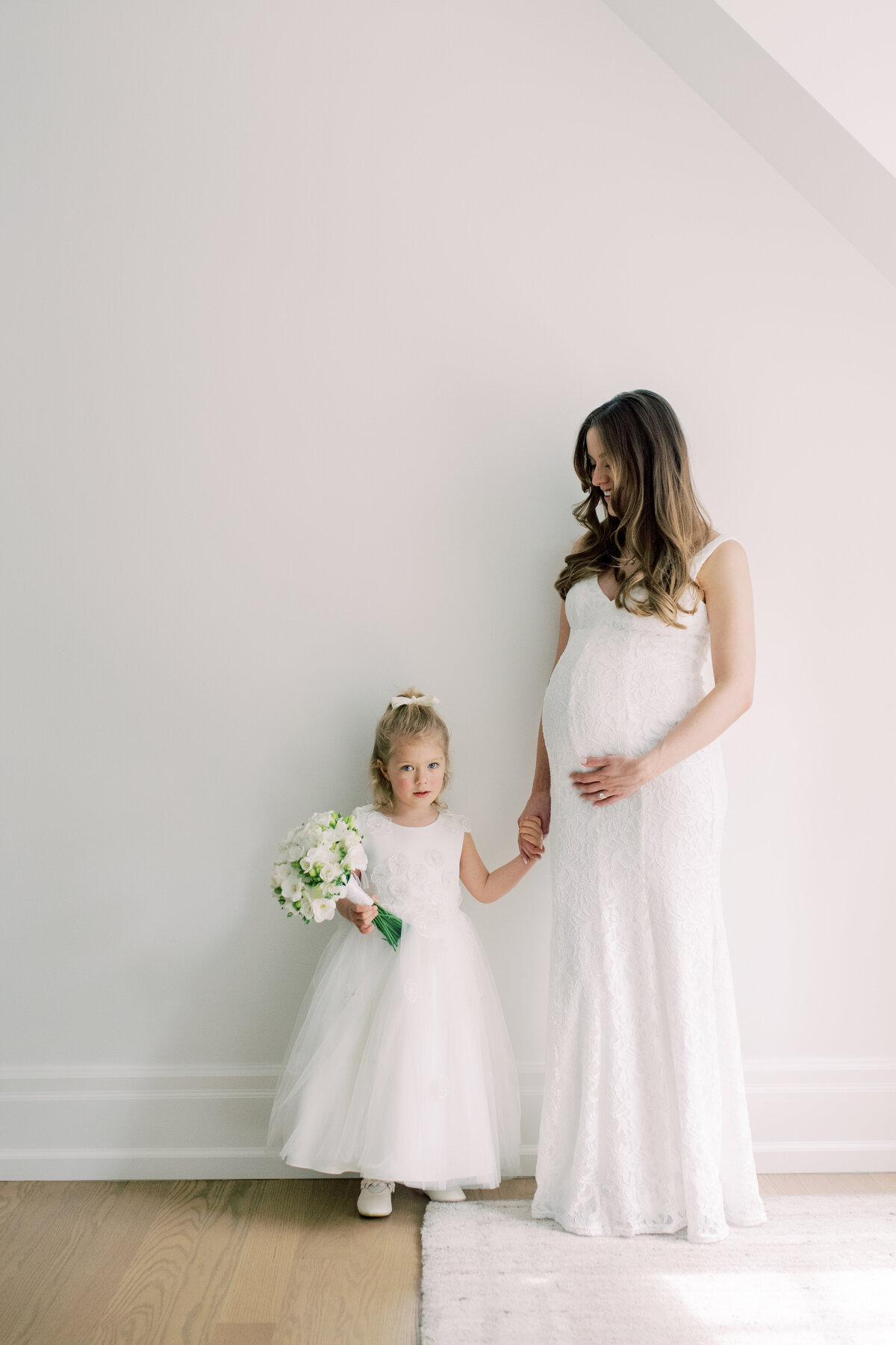 A sweet image of a pregnant bride holding hands with a young flower girl, both dressed in white, sharing a gentle moment together