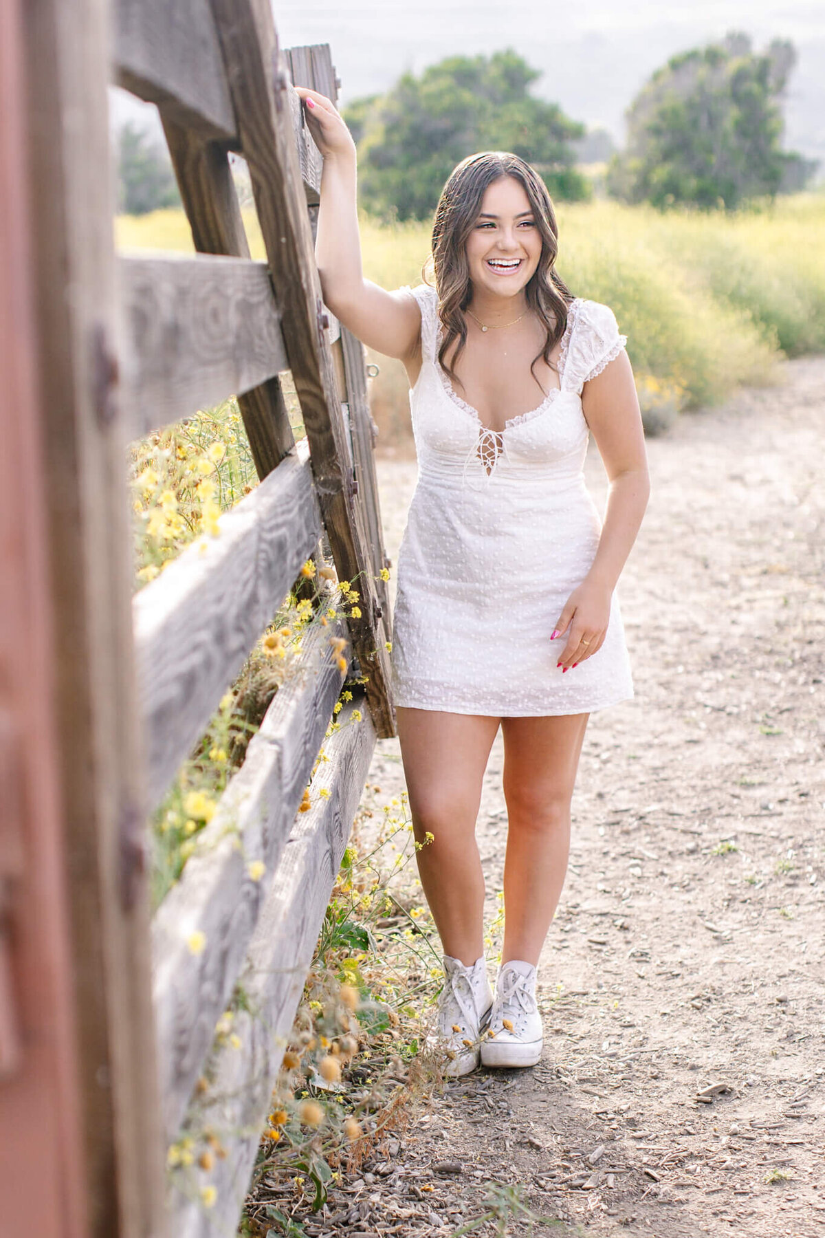 A woman in a white dress and sneakers leans on a wooden fence, smiling in a sunny outdoor setting with greenery in the background.