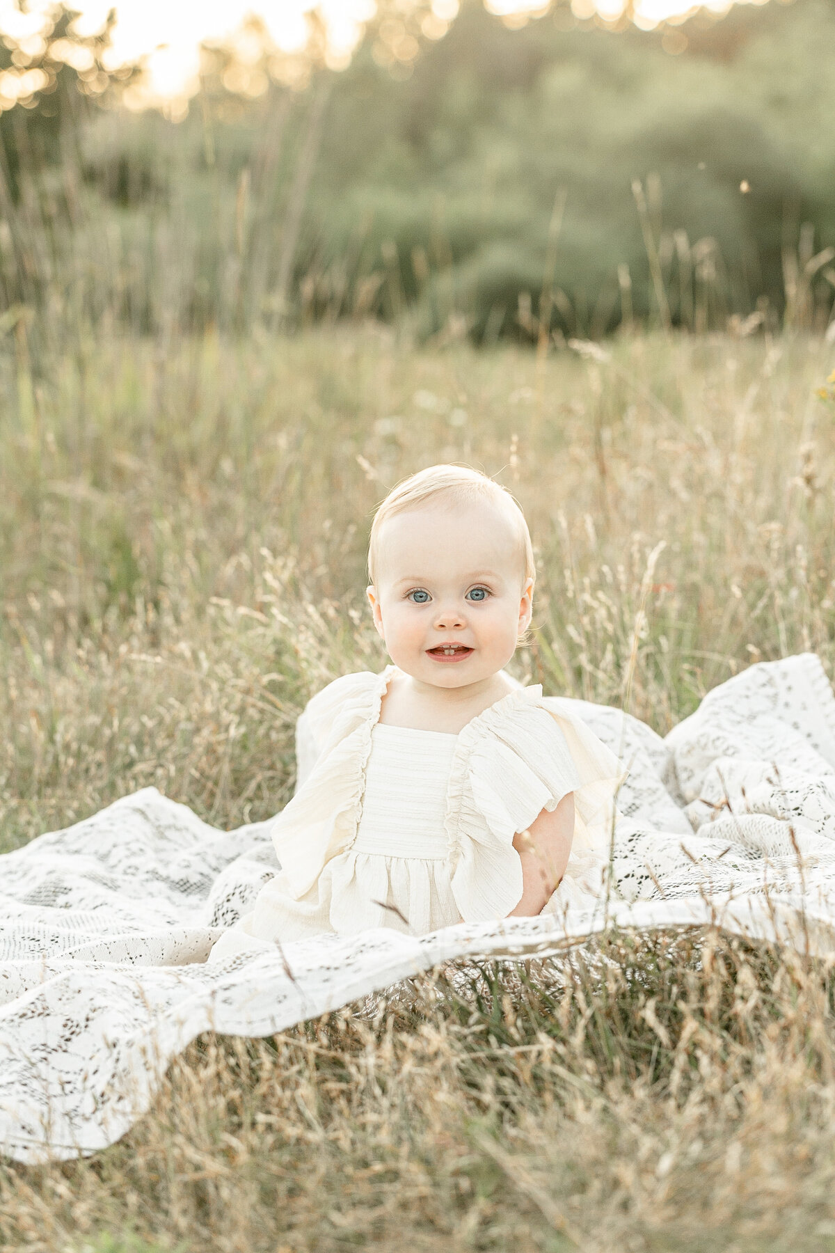 One year old light skinned girl in ivory dress sitting on a lace blanket outdoors surrounded by tall golden grasses. Outdoor sunset session in Portland oregon.