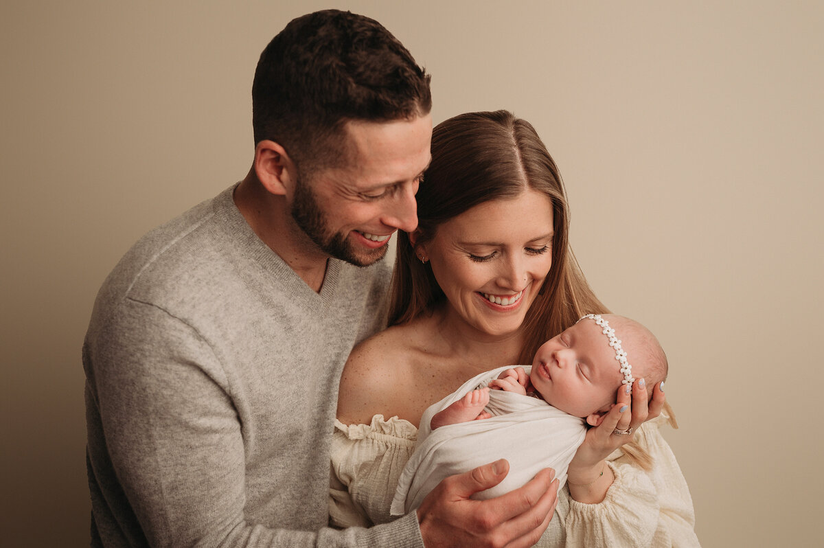 smiling New parents looking at and holding their brand new baby girl who is swaddled in a white swaddle with toes peeking out and white headband