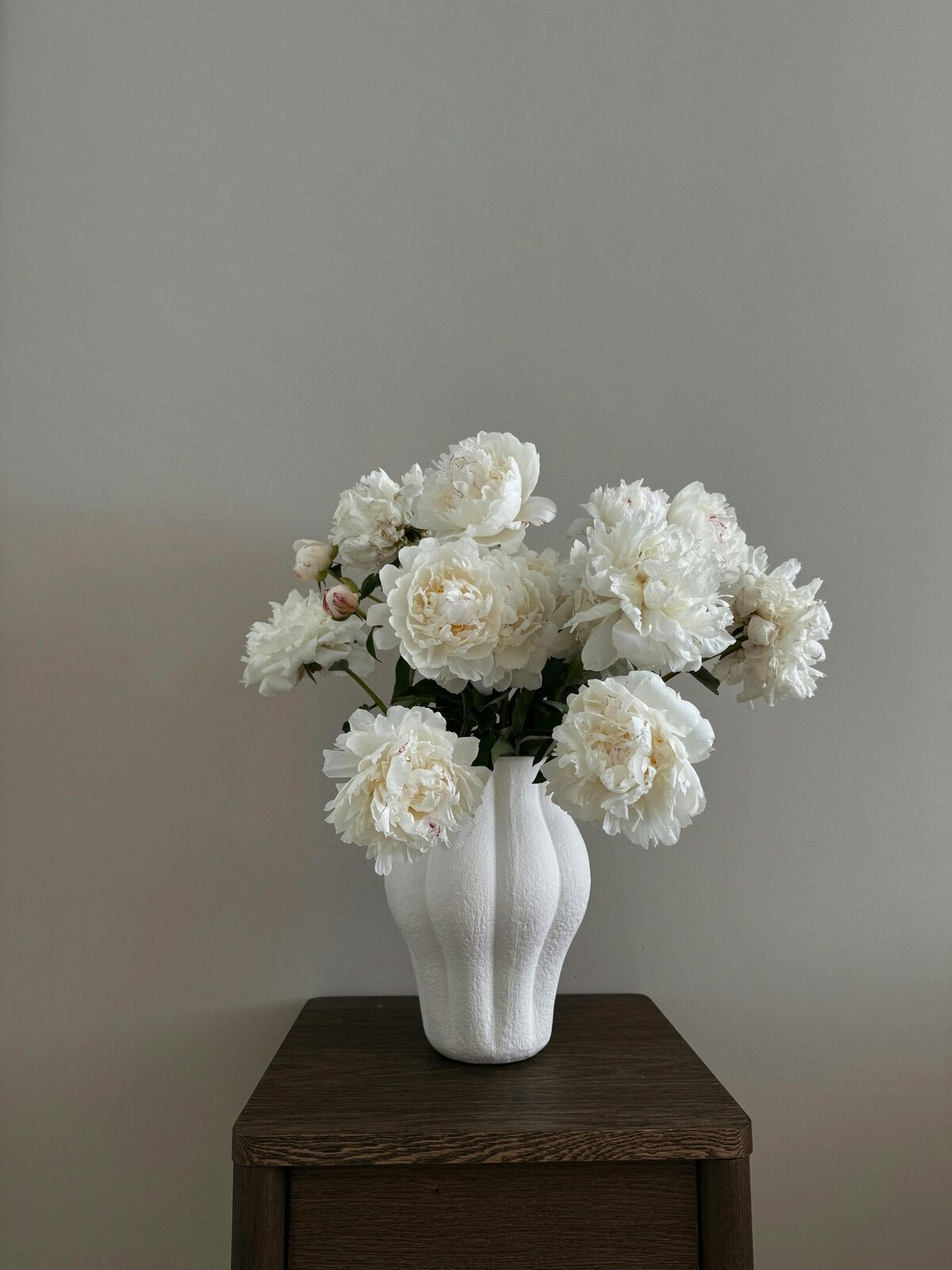 A textured white vase filled with full white peonies in bloom, elegantly placed on a small walnut side table.