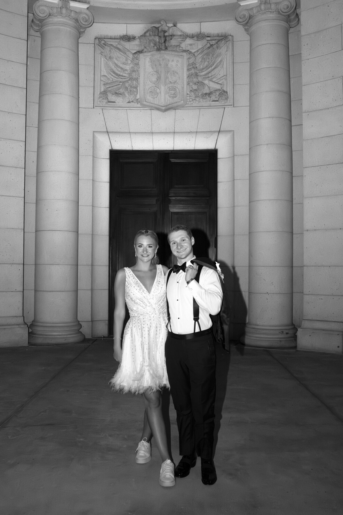 A couple in formal attire stands smiling in front of a large wooden door. The woman wears a short dress and sneakers, while the man is in a suit and bow tie. They are framed by ornate pillars and a carved stone relief above. The image is in black and white.