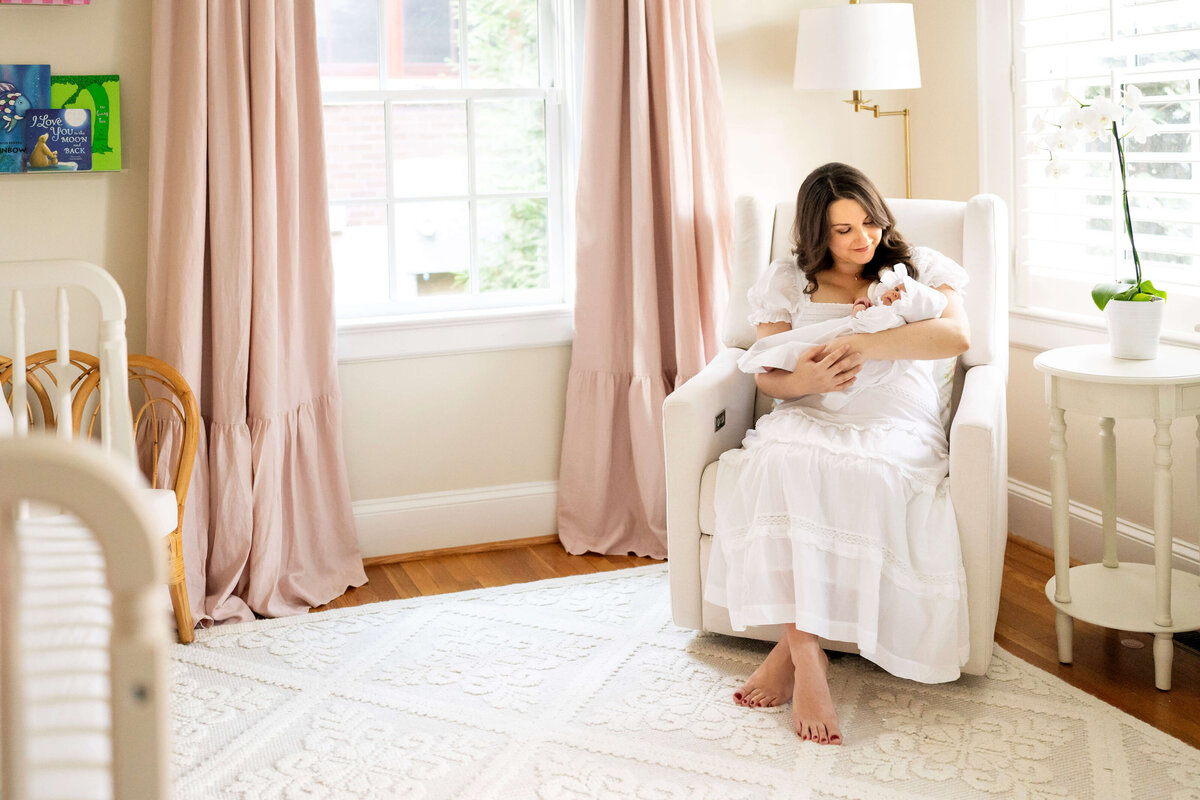 Mother in white dress sitting on rocking chair and rocking newborn girl