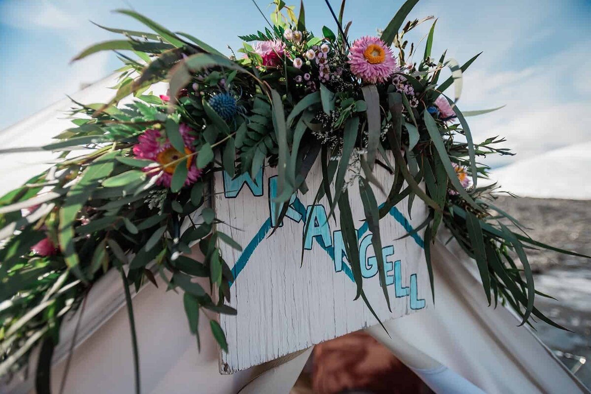 a light colored canvas bell tent on the Matanuska Glacier in Alaska is decorated with wedding flowers as enhanced decor over the arch of the doorway during a private luxury helicopter glacier elopement. In the sky behind the flowers that fill the frame is a blue sky with wispy white clouds and mounds of ice and snow.   The flowers partially obscure a tent name placard that says Wrangell in painted blue font, an unusual details for a luxury elopement on a glacier.