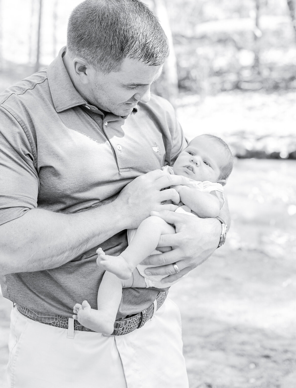 Newborn baby in father's arms – captured by a Birmingham newborn photographer. A sweet newborn gazes softly while cradled in dad’s strong hands, capturing the warmth and security of early parenthood in this natural, outdoor session.