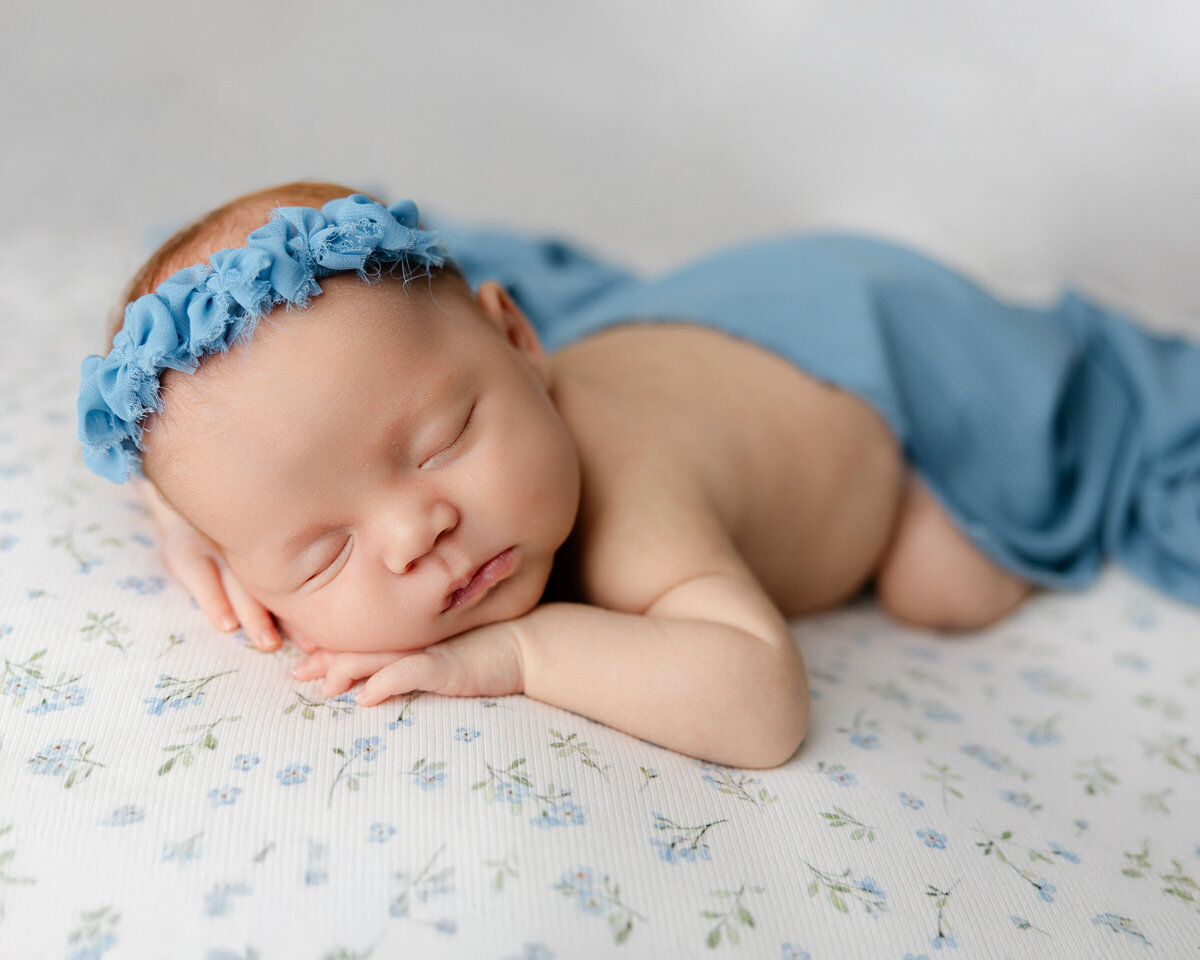 Newborn baby boy peacefully sleeping on a soft, floral-themed backdrop, wrapped in a cozy blue blanket with a matching flower headband. Captured during his newborn session by Yucaipa newborn photographer Kaitlyn Dawn Photography.