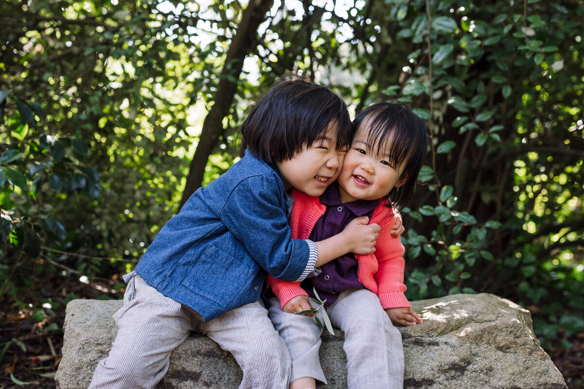 4-year-old boy sitting on rock and hugging toddler sister sitting next to him and both are smiling in San Francisco garden
