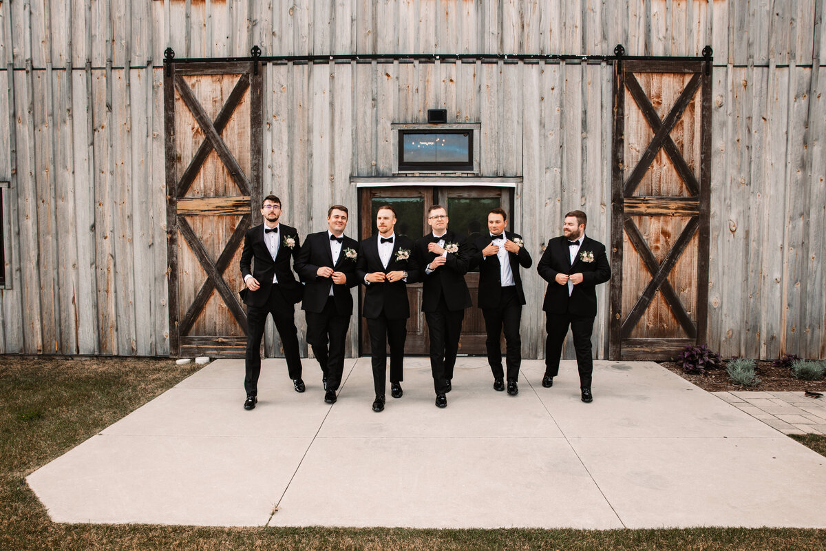 A groom adjusts his cuffs while walking in front of a barn with his groomsmen