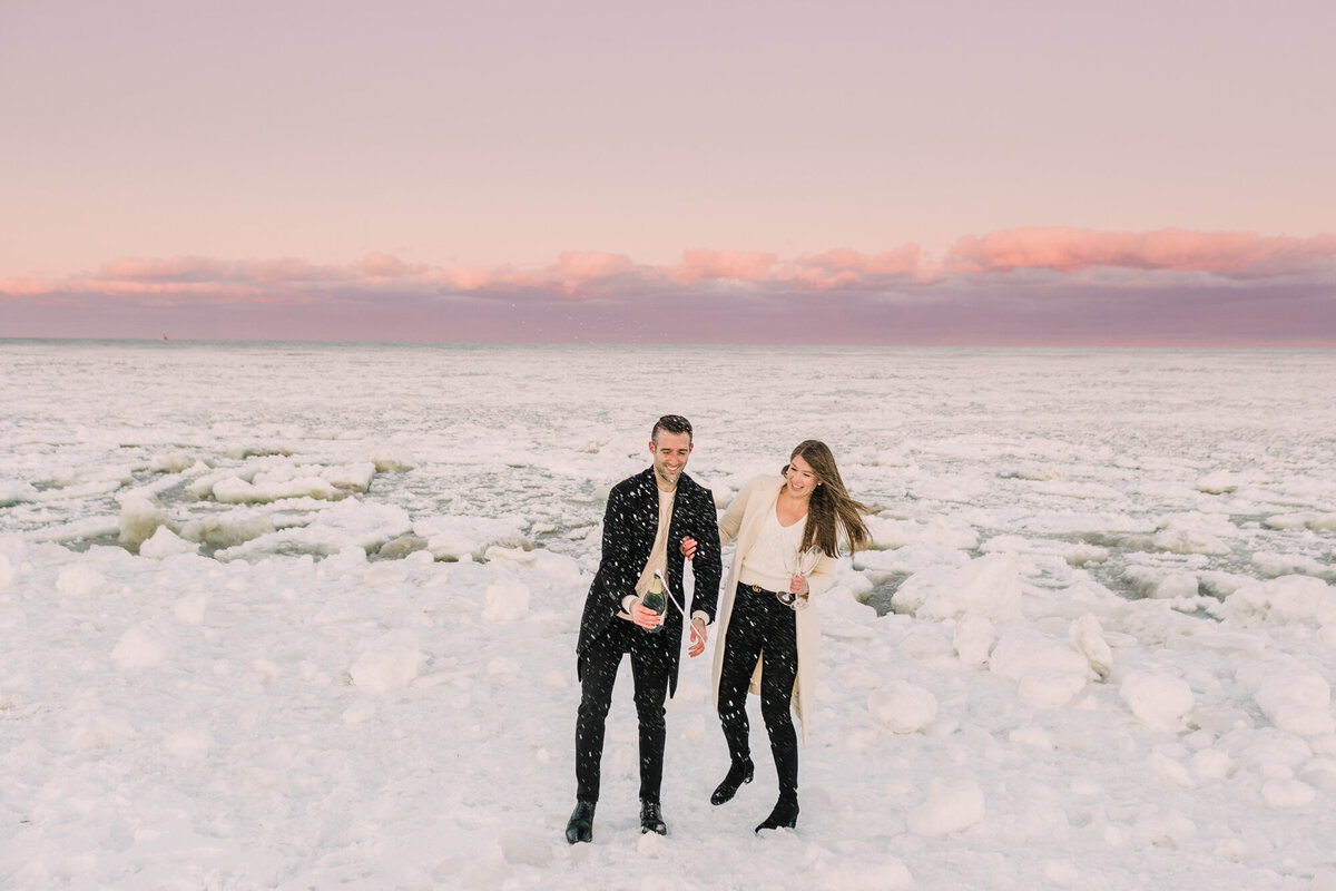 A couple opens a bottle of champagne along a frozen Chicago lakefront for an engagement photo
