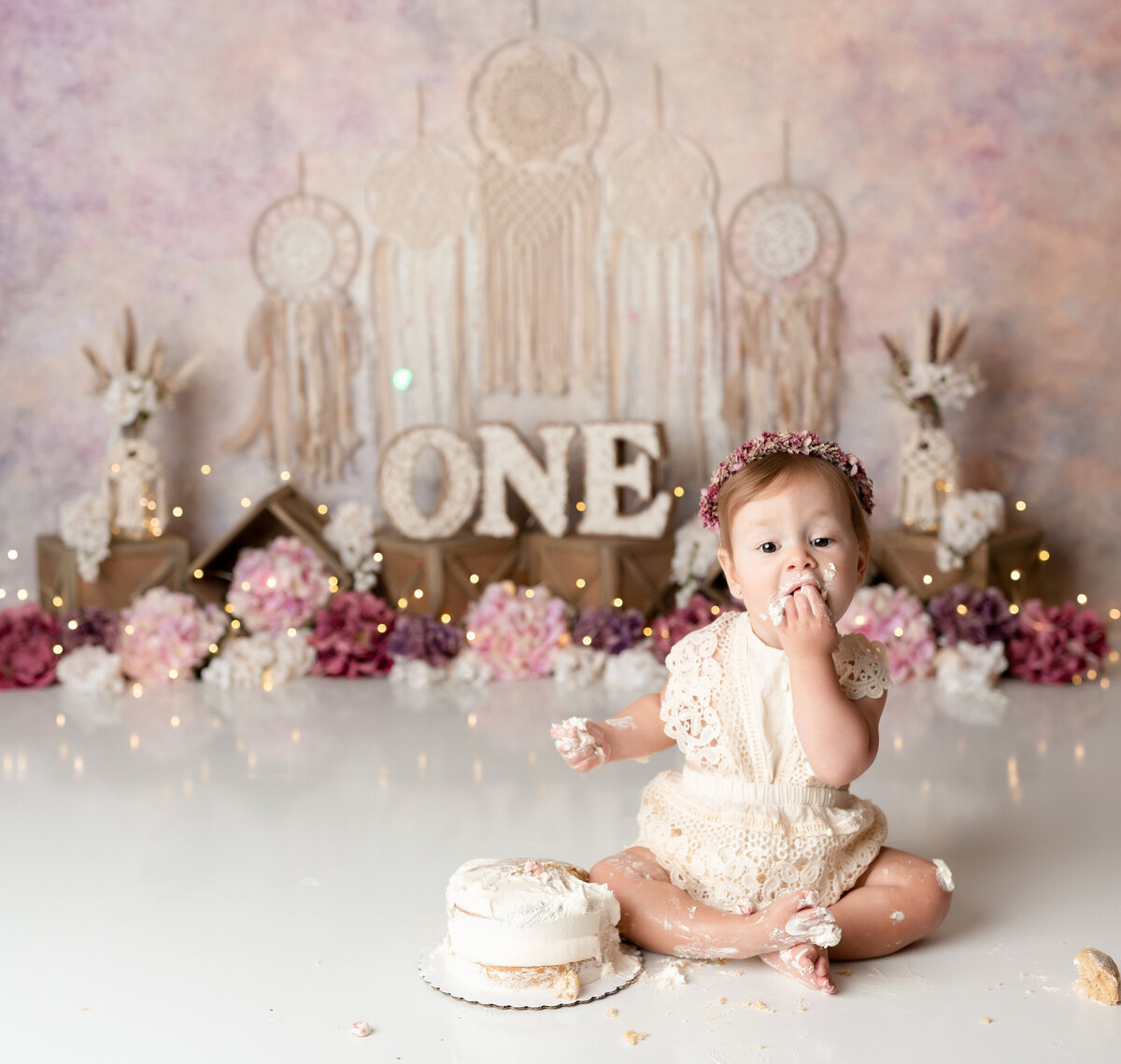 Boho themed cake smash in West Palm Beach, FL newborn and cake smash photography studio. Baby girl in cream lace outfit is eating a fist full of cake. In the background there are macrame wall hangings, flowers, and subtle twinkle lights.