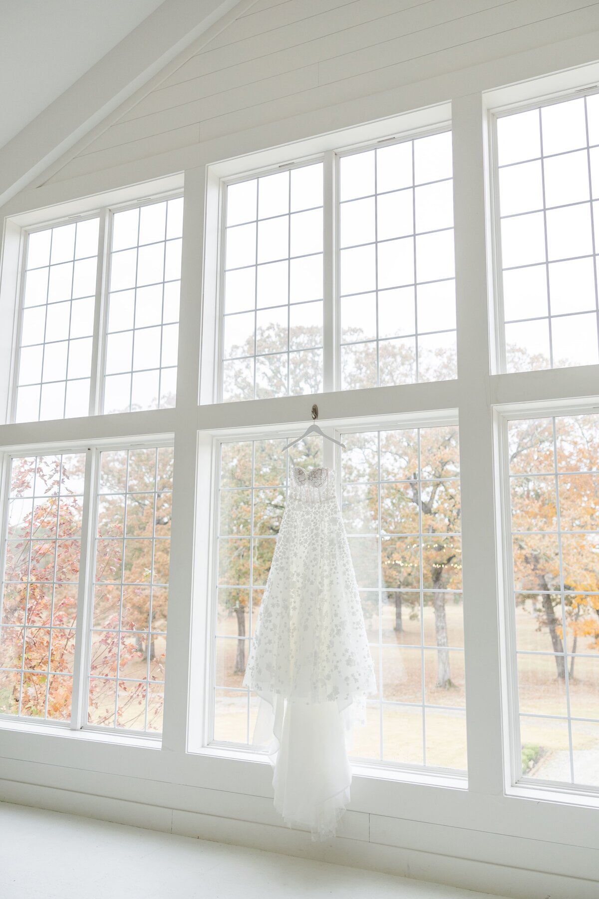 A white lace wedding gown hangs on a wall of windows taken by an Arkansas wedding photographer