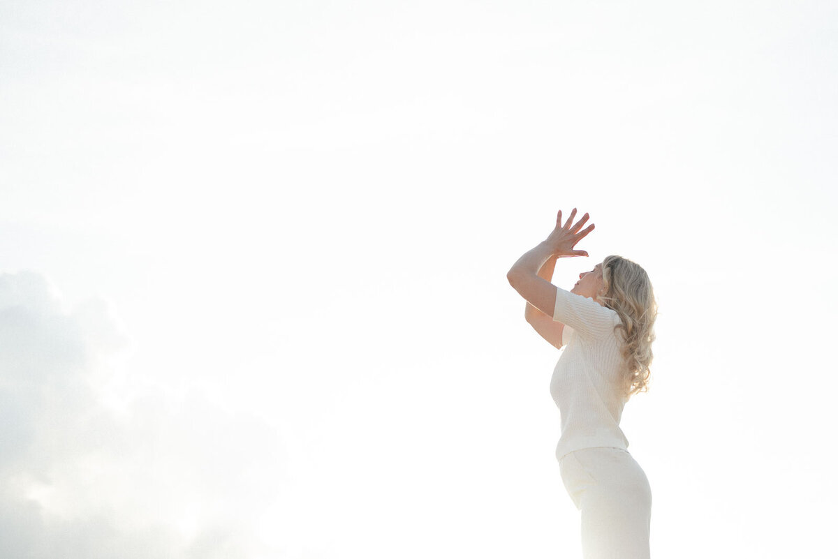 Eine Healerin steht in weißer Kleidung mit gefalteten Händen in der Natur und die Kamera zeigt Richtung Himmel