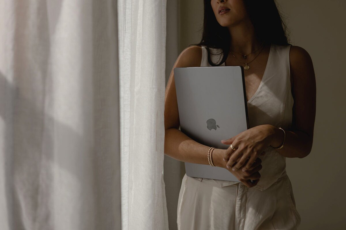 A woman in a beige linen vest and matching trousers, accented with gold jewellery, holds a laptop to her chest as she stands in quiet reflection. Soft light filters through white linen curtains, framing the moment with an air of understated elegance.