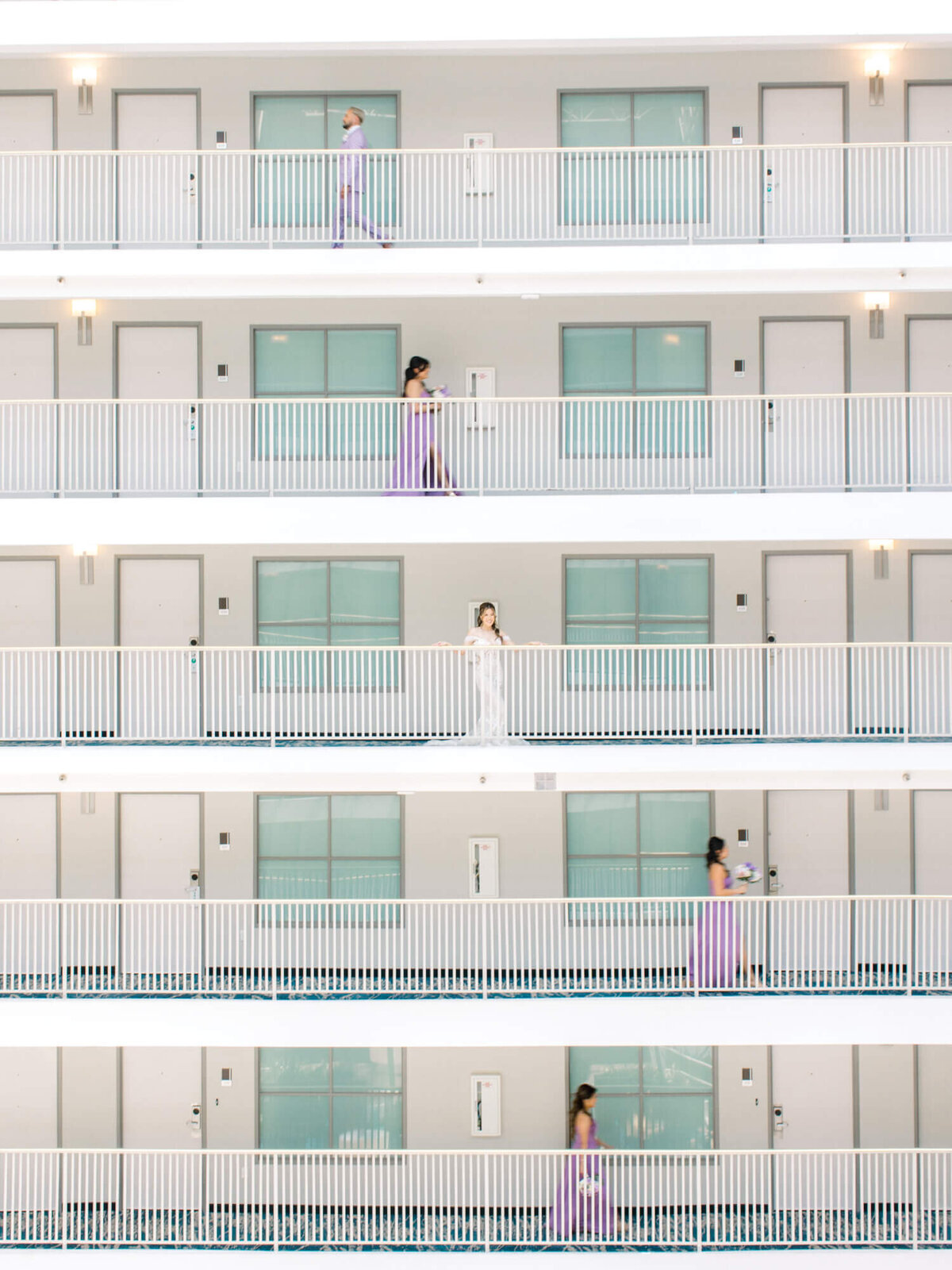 Five bridesmaids in purple attire walk along different levels of a white building with turquoise doors. The scene conveys a lively, dynamic feel.