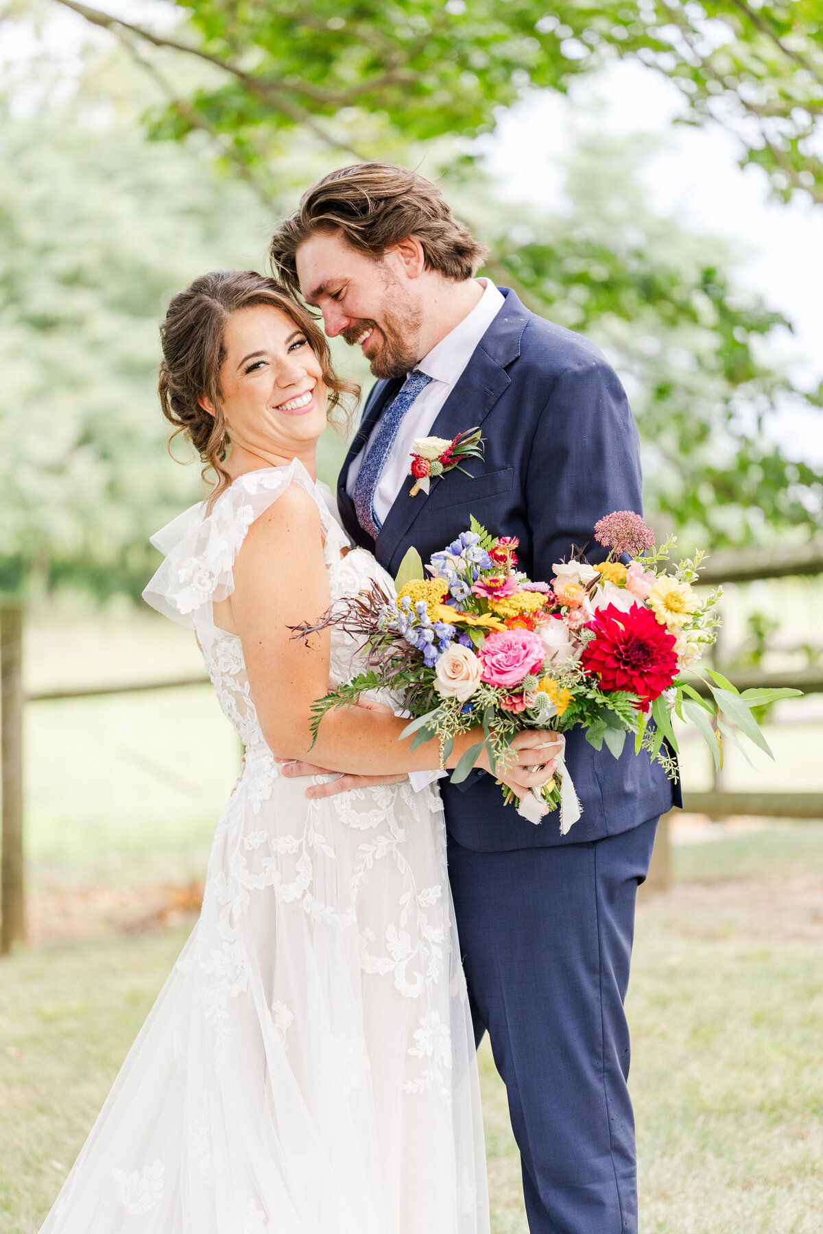 A bride and groom in a lace dress and blue suit laugh while snuggling in a field with a colorful bouquet held in front of them
