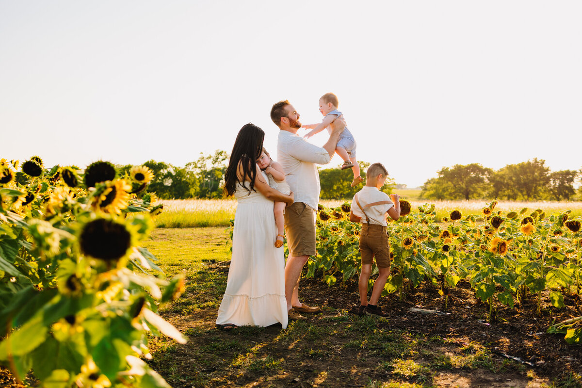 The family poses for a professional spring mini photo shoot. Mom is wearing a long white dress and dad is wearing brown pants and white t-shirt. Around them are yellow daisies and the children play with them.