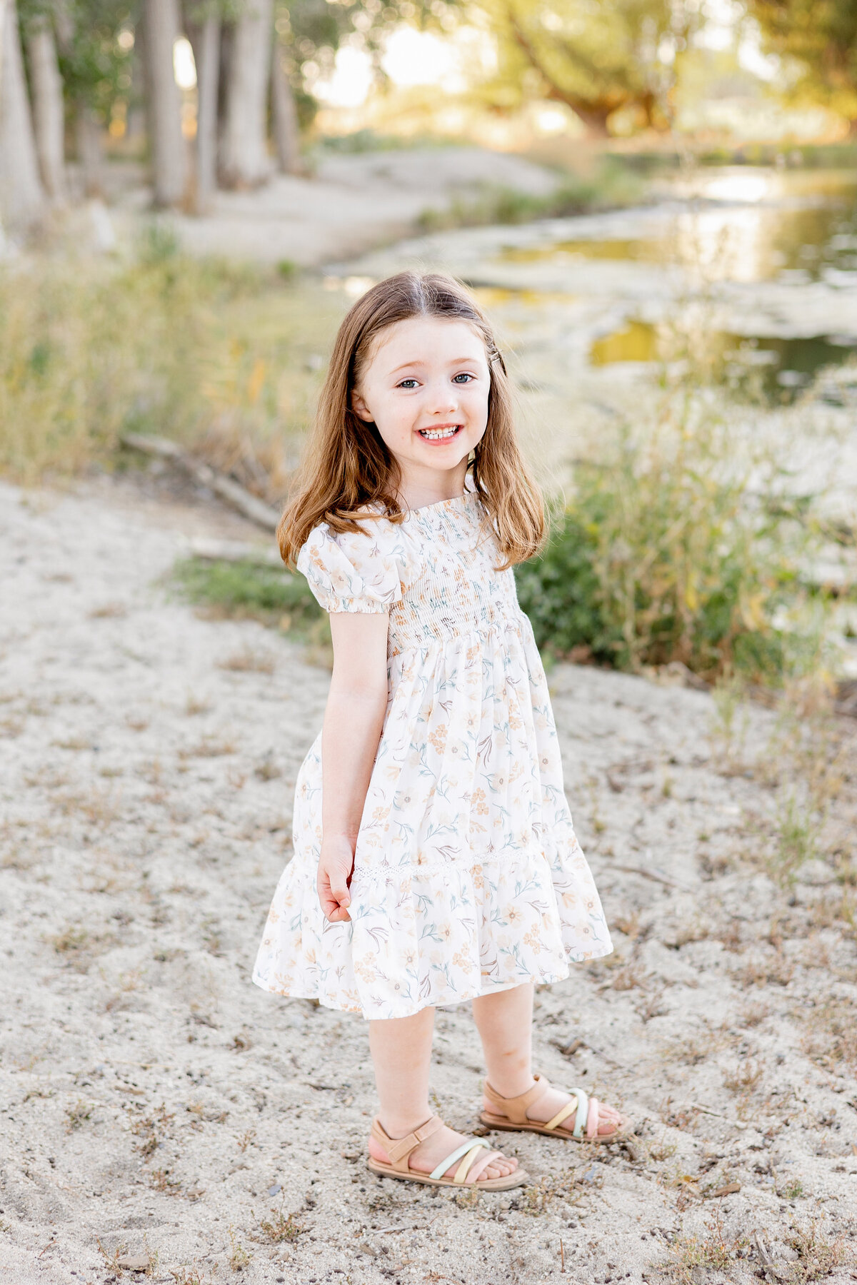 A young girl standing near a peaceful lake, smiling brightly while wearing a floral dress.
