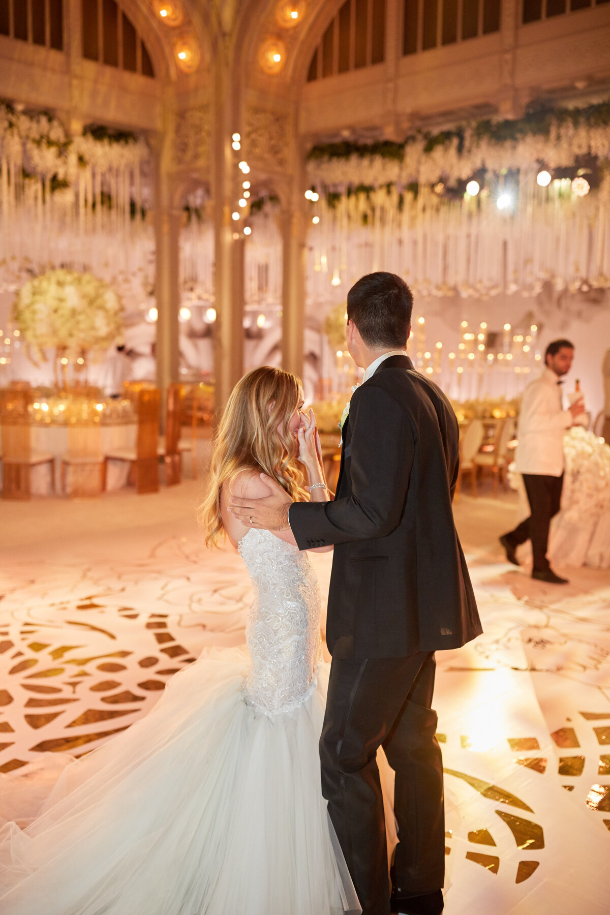 The beautiful moment captured in the photograph took place at the iconic New York City location, The New York Public Library, as documented by the talented photographer Christian Oth, with videography by East West Films. In the scene, a couple is seen sharing a dance at their wedding reception within an elegant and warmly lit venue. The bride, donning a white strapless gown, delicately holds her hand up to her face while the groom, dressed in a dapper black suit, faces away from the camera. The venue is adorned with a charming display of flowers and twinkling string lights, adding to the romantic ambiance of the occasion.