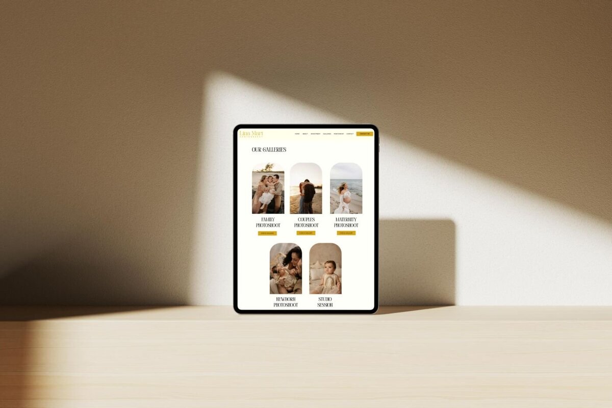 Tablet standing upright on a wooden surface in soft sunlight, showing a photography website gallery page featuring five categories: family, couples, maternity, newborn, and studio sessions with warm lifestyle portraits.