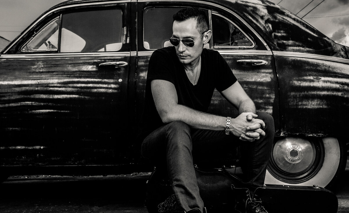 Male musician photo Adam Rutledge black and white wearing black cloths sitting against old black car