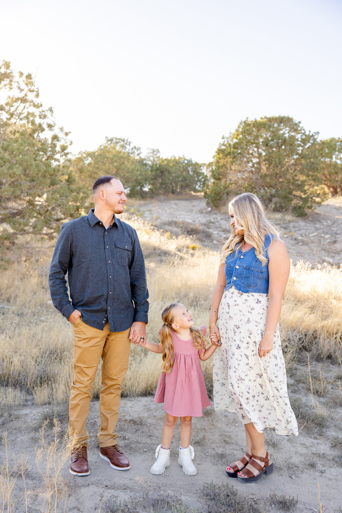 A couple and their young daughter hold hands while walking through a scenic, dry landscape in Eagle Mountain, Utah. The little girl in a pink dress looks up at her parents, who are smiling at each other in the golden sunlight.